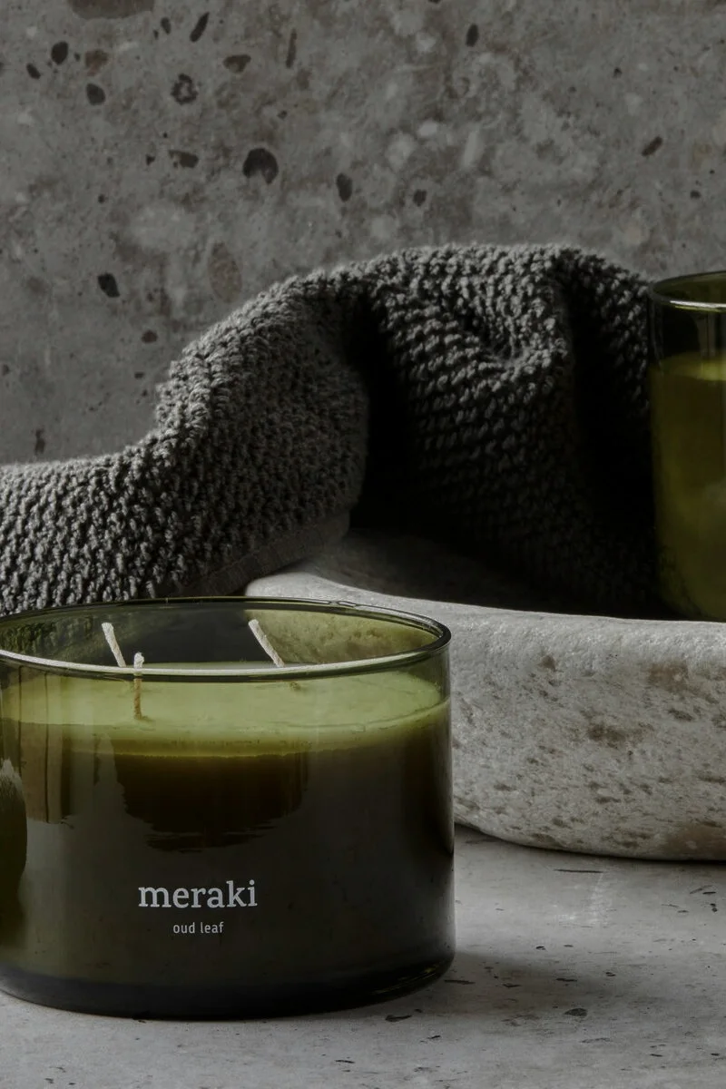 Dark green glass candle with "meraki oud leaf" label, green wax, and two wicks, next to a grey towel and stone bowl.