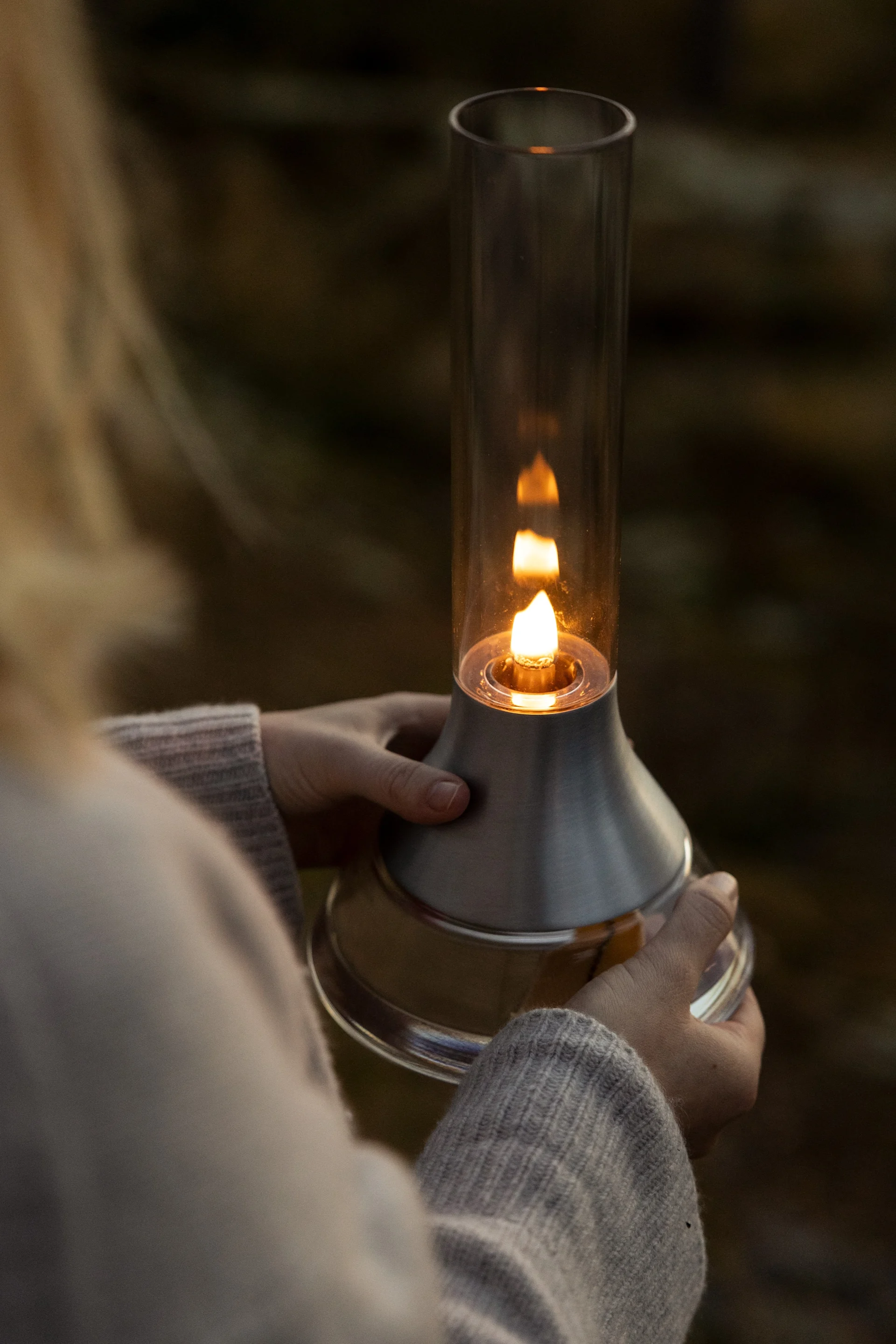 Person holding a modern lantern with a lit flame in a glass cylinder and brushed metal base.