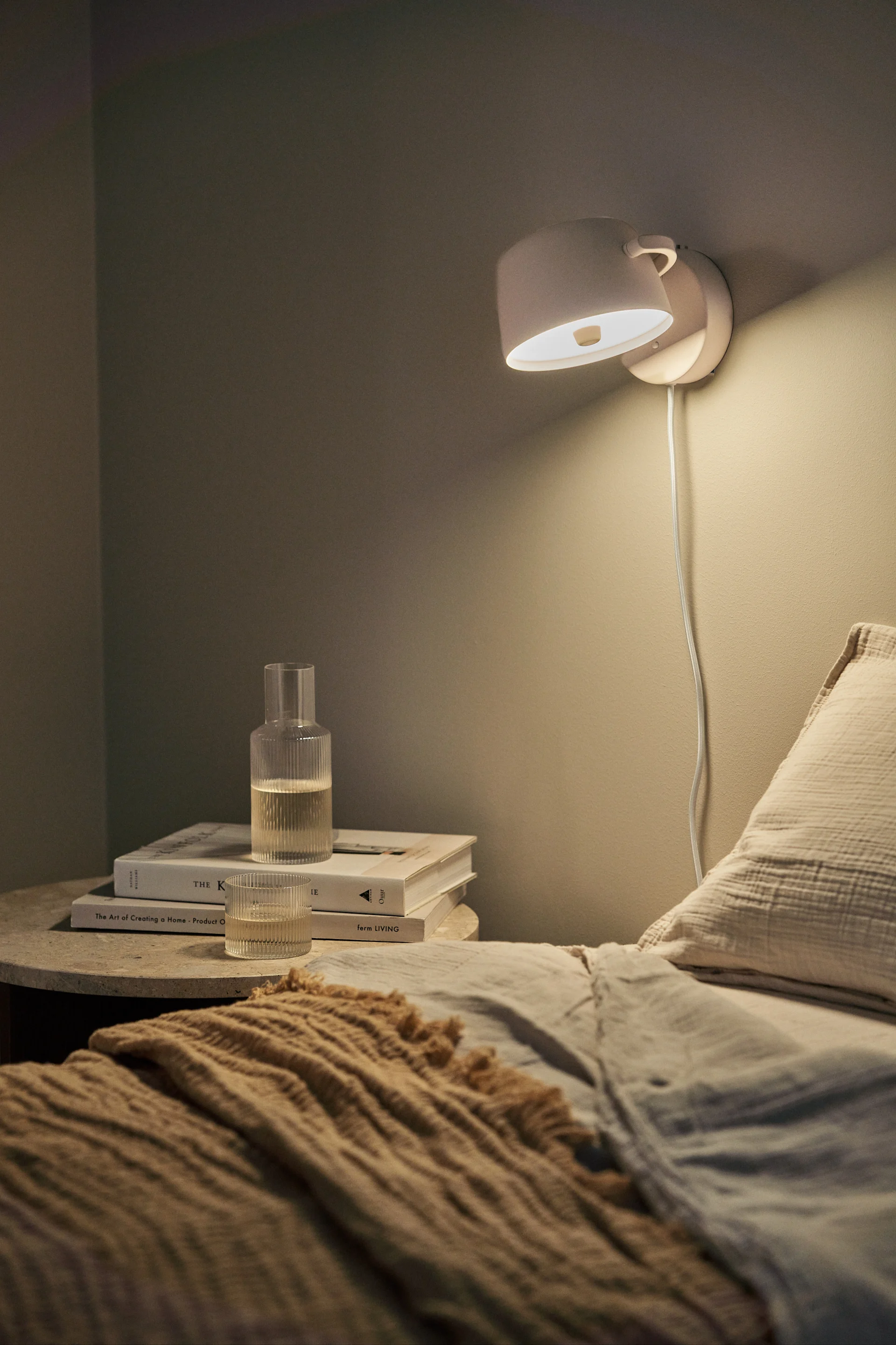 A wall lamp illuminates a cozy bedroom, featuring a bed with linen and a knit throw, and a side table with books and glass drinkware.
