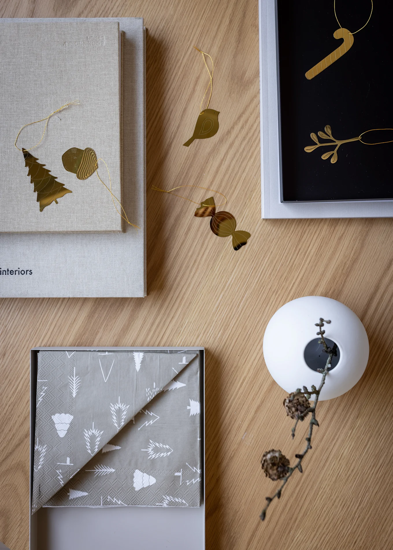 A flat lay shows gold Christmas ornaments (tree, acorn, bird, candy cane, candy), grey patterned napkins, and a white vase with a branch on wood.