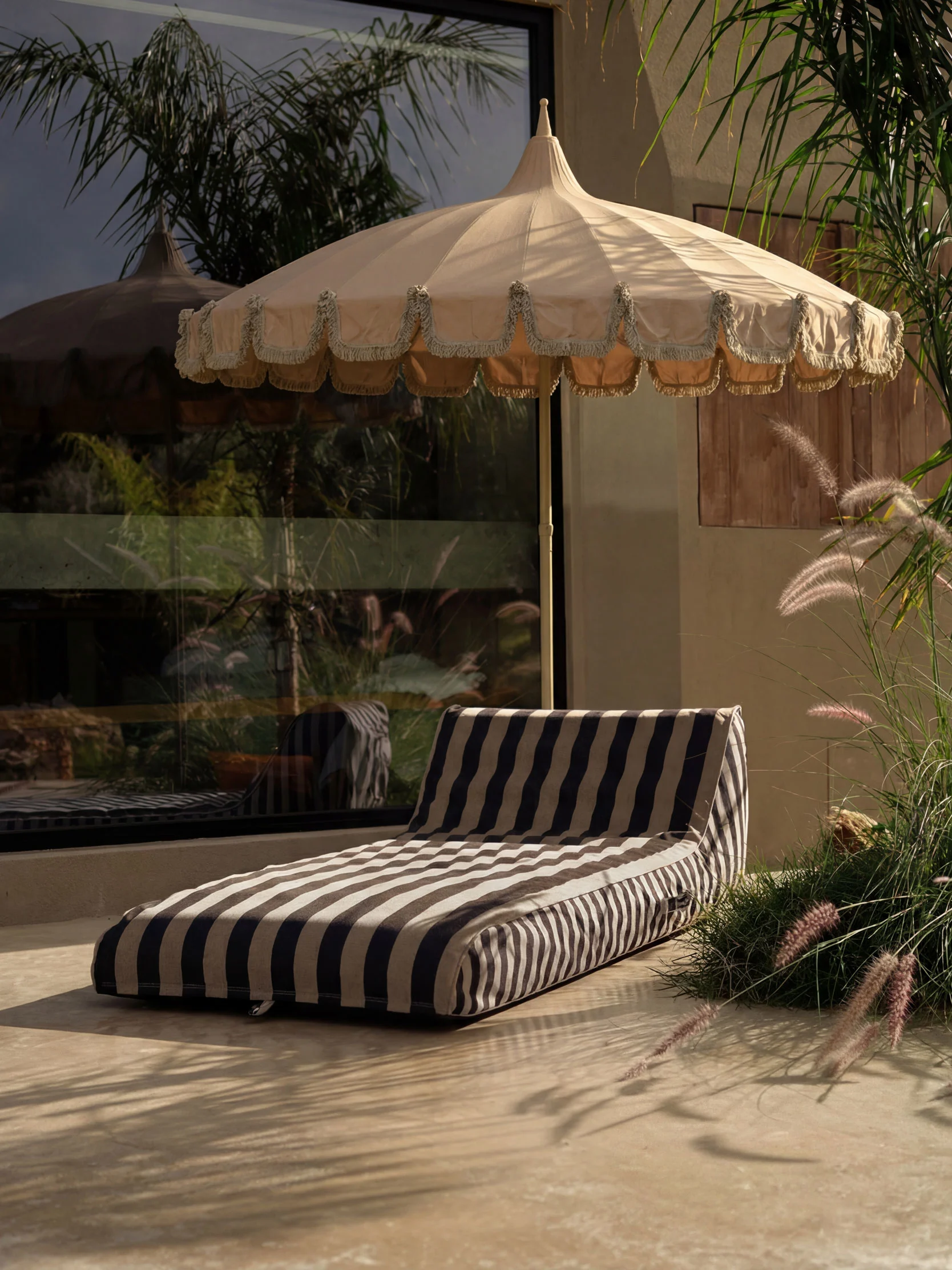 Outdoor patio with a beige fringed parasol and a striped lounge cushion on light flooring, surrounded by lush plants.