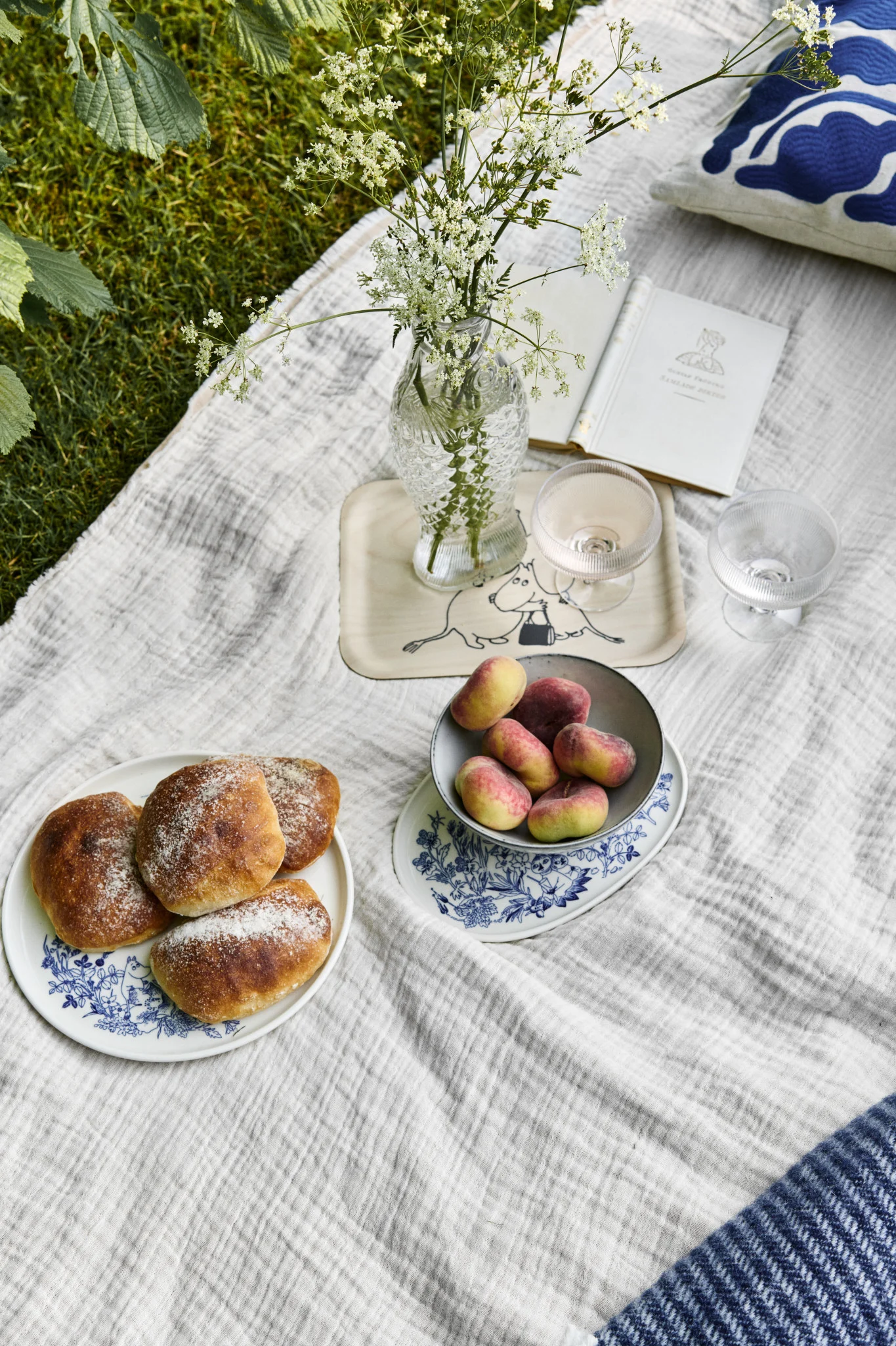 Picnic spread on grass with buns, flat peaches, wildflowers in a vase, and an open book on a white blanket.