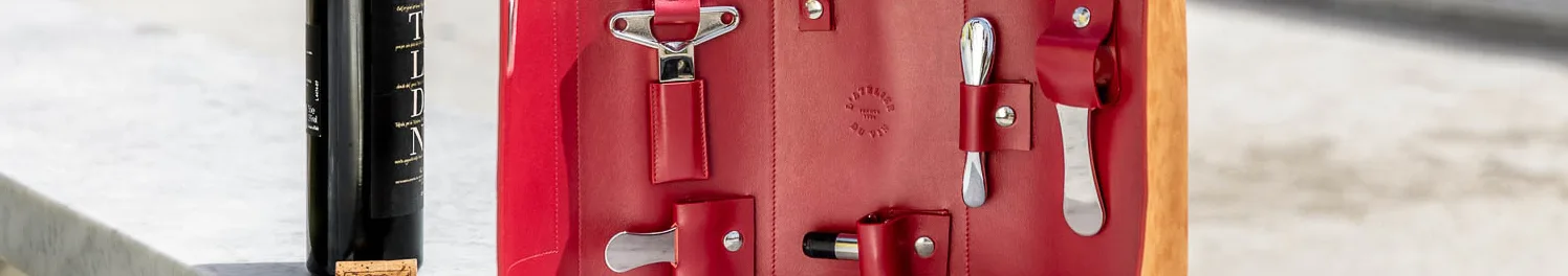 A dark wine bottle stands beside an open red leather roll with silver wine tools secured inside.
