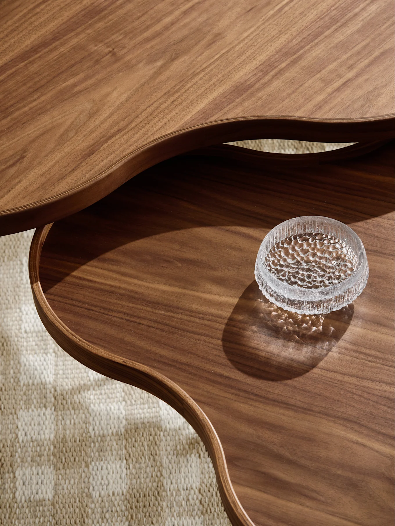 Close-up of a multi-tiered wooden coffee table with wavy edges and a textured glass bowl on the lower tier, on a textured rug.