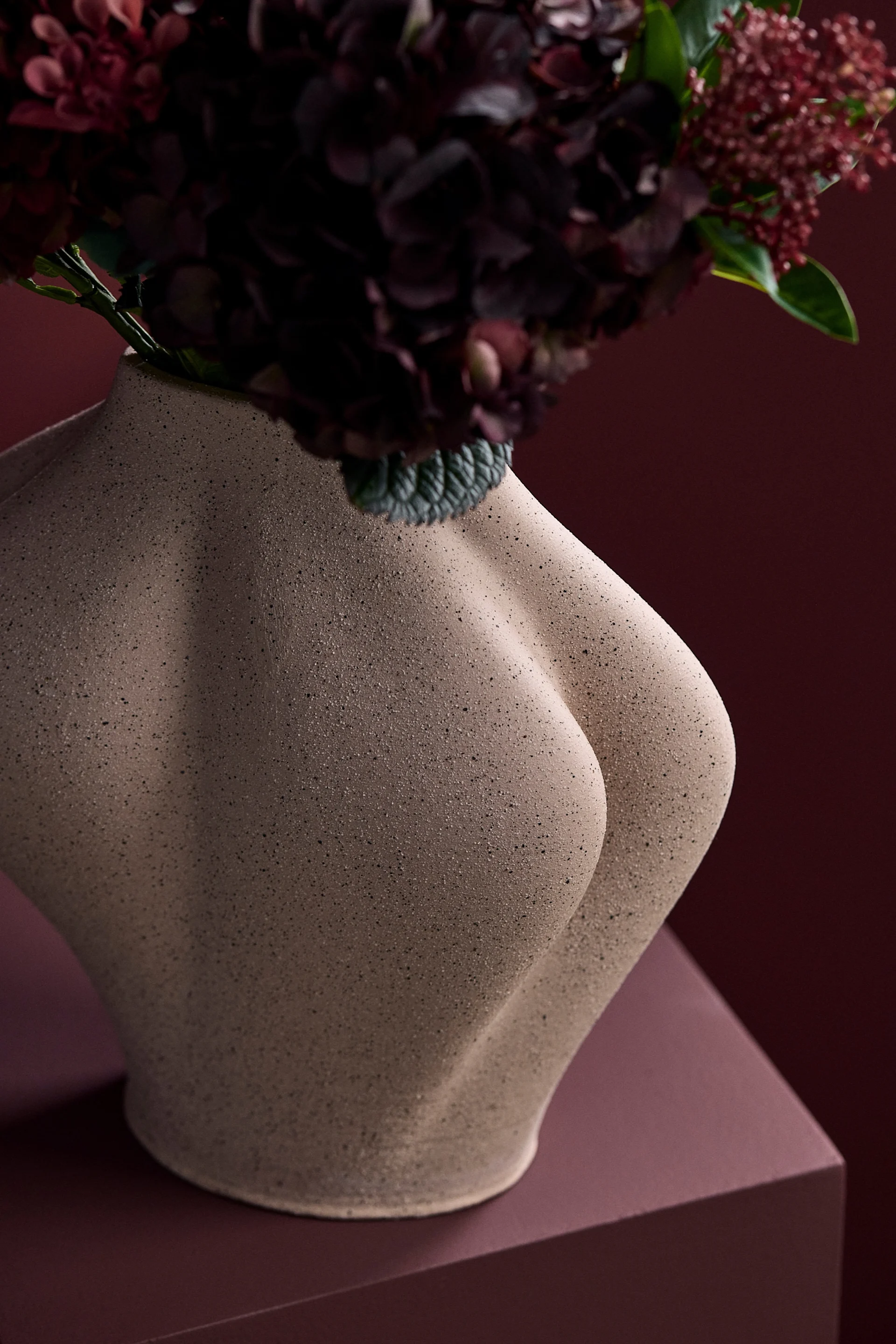 Sculptural beige vase shaped like a human torso, holding dark red flowers against a matching red background.