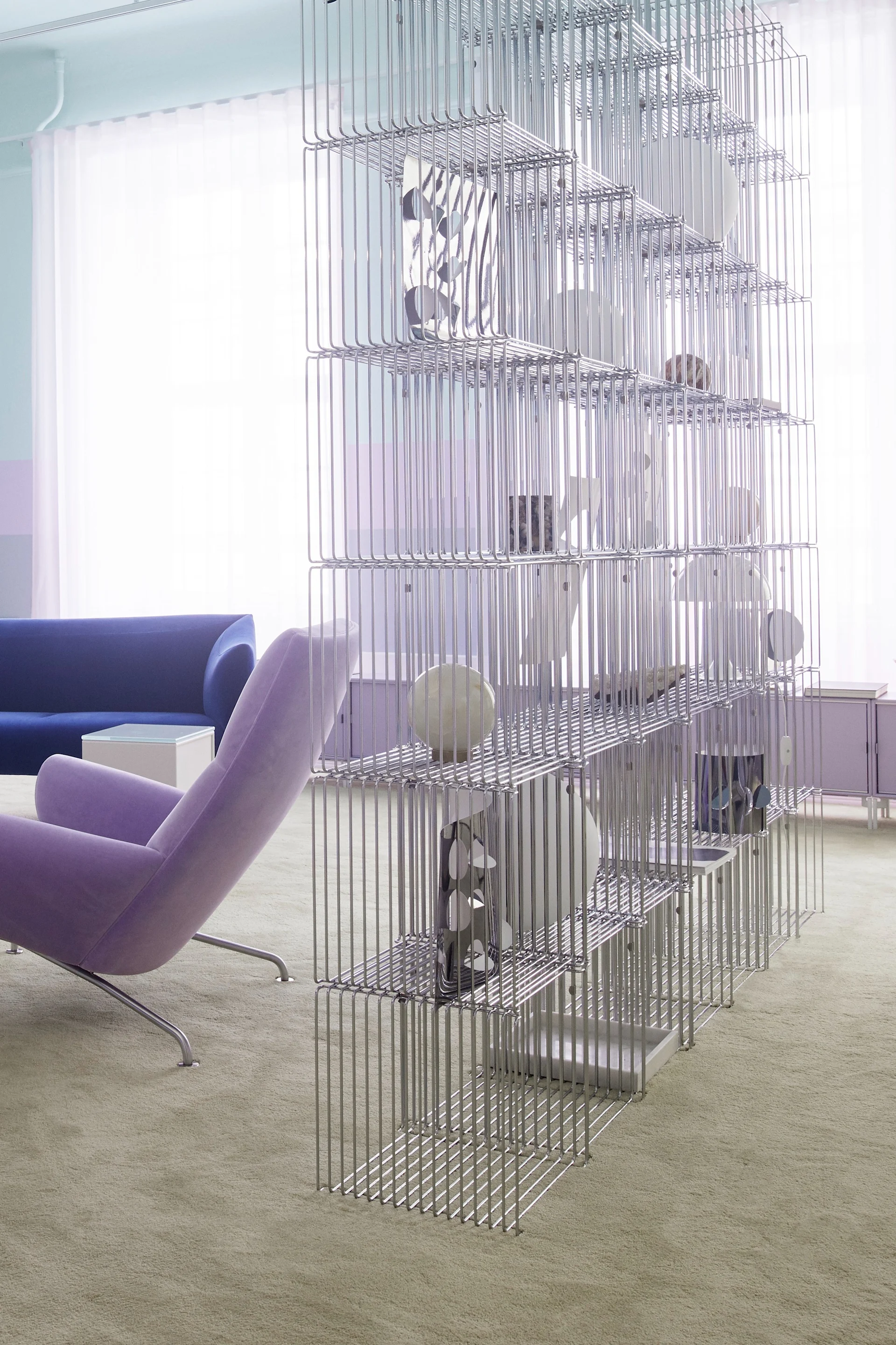 Modern room with a tall, chrome-plated metal wire shelving unit, a purple armchair, and a blue sofa on a light green carpet.