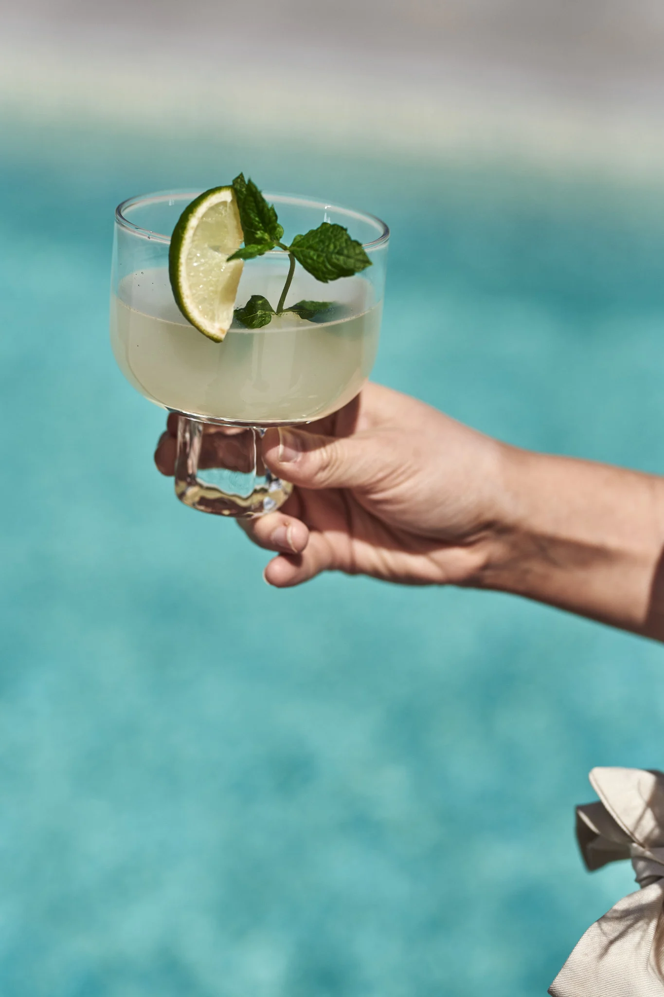 Discover these refreshingly simple summer cocktails and mocktails. Here you see a hand hold a mojito with a wedge of lime next to a pool. 