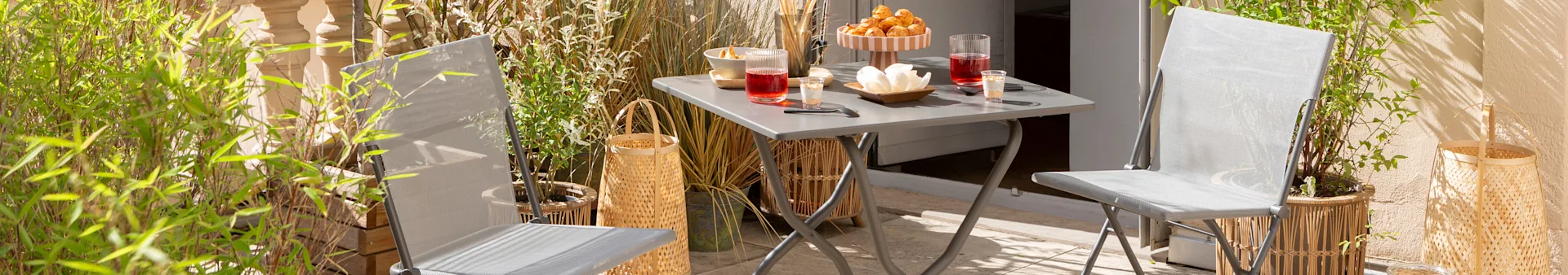 An outdoor patio with two grey garden chairs, a matching folding table with drinks and snacks, and various potted plants.