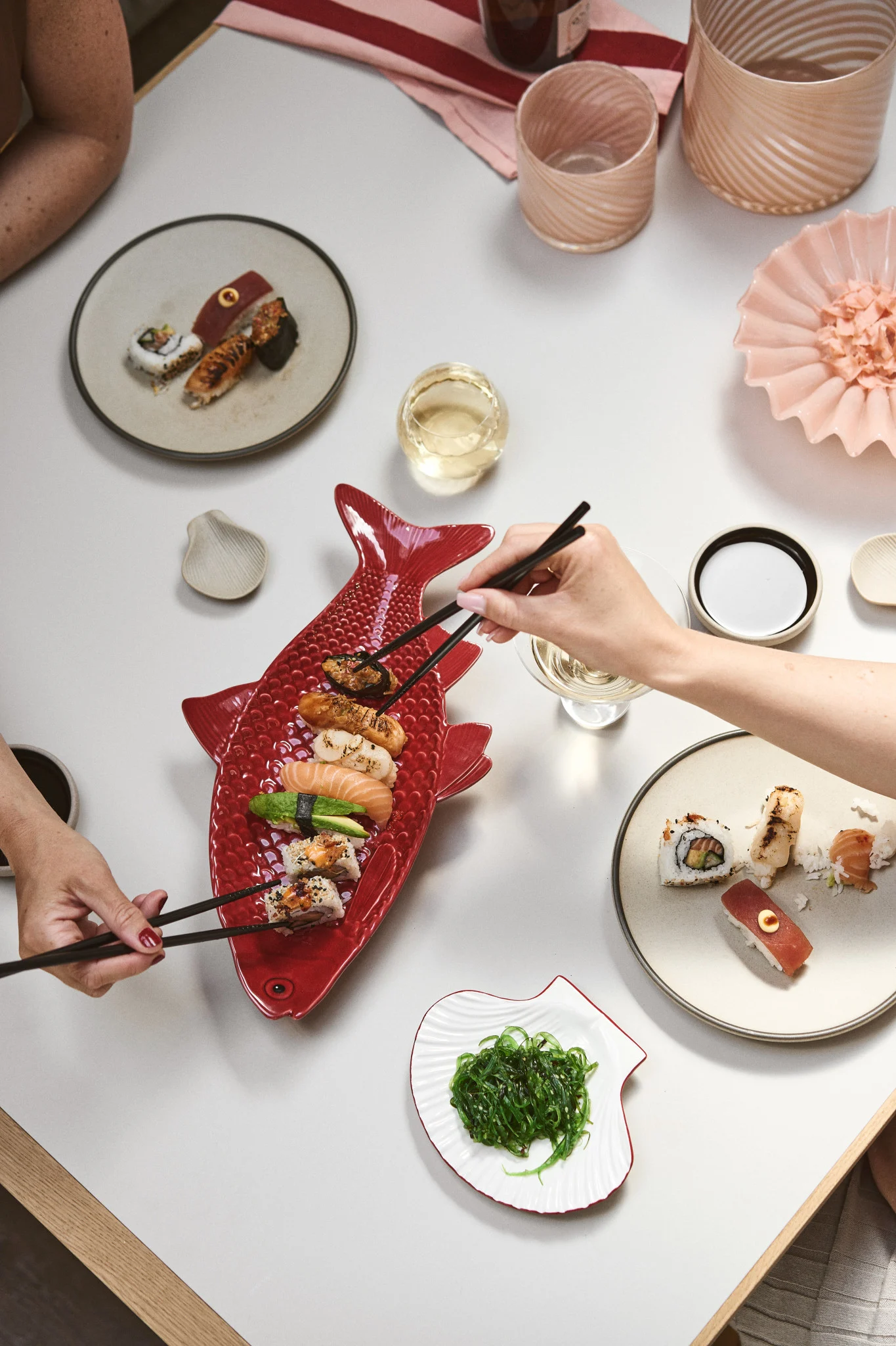 Two people sharing a large red fish-shaped platter of sushi with chopsticks, alongside other sushi plates, wine, and seaweed salad.