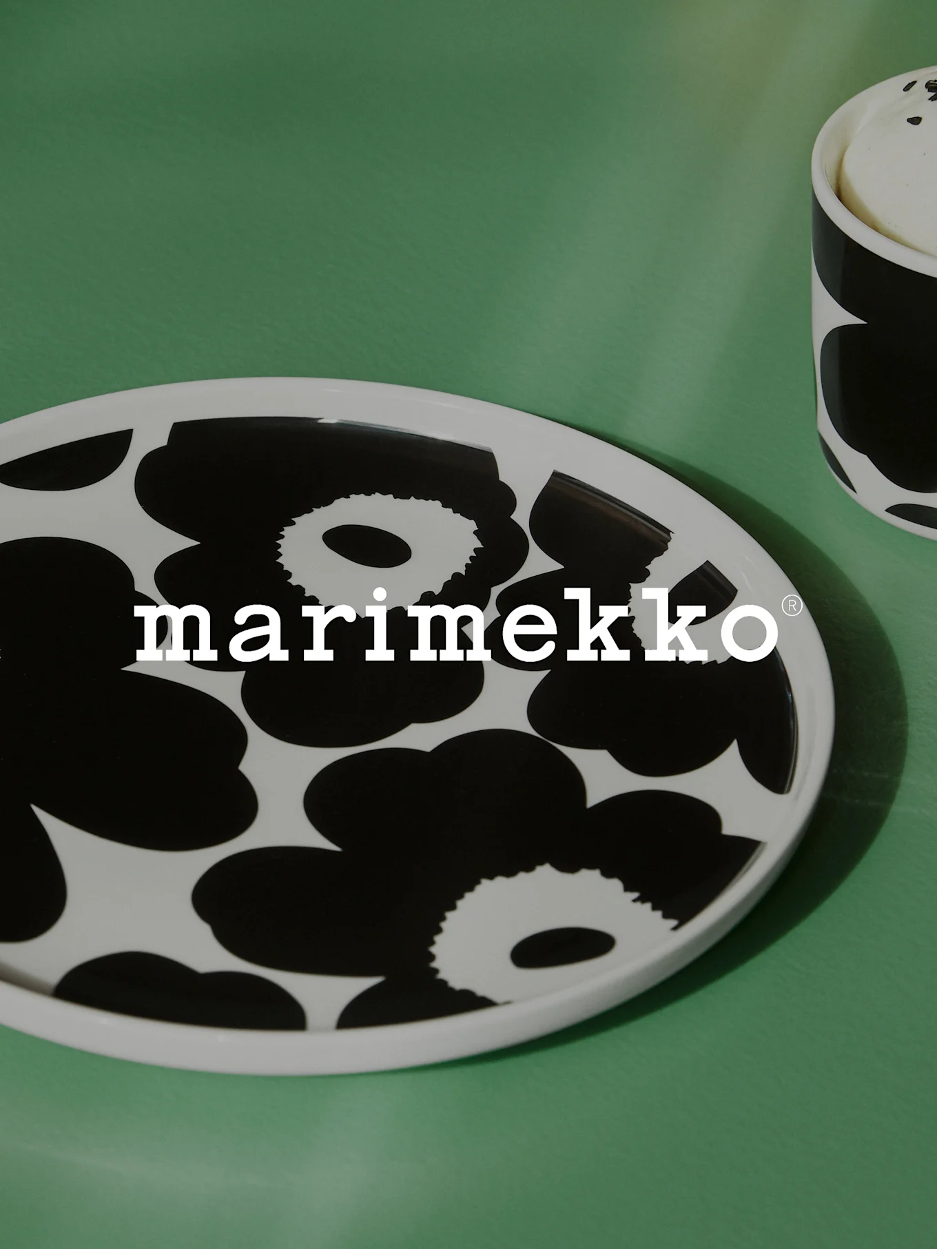 White oval plate with black floral pattern and Marimekko logo on a green surface, with a partial matching mug.