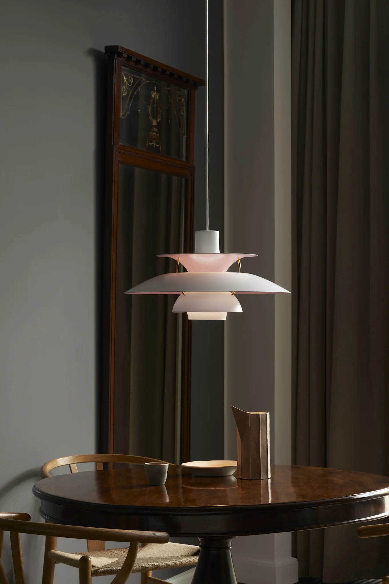 A multi-tiered pendant light with white and pink shades hangs over a round, dark wooden table. A decorative mirror and curtains are in the background.