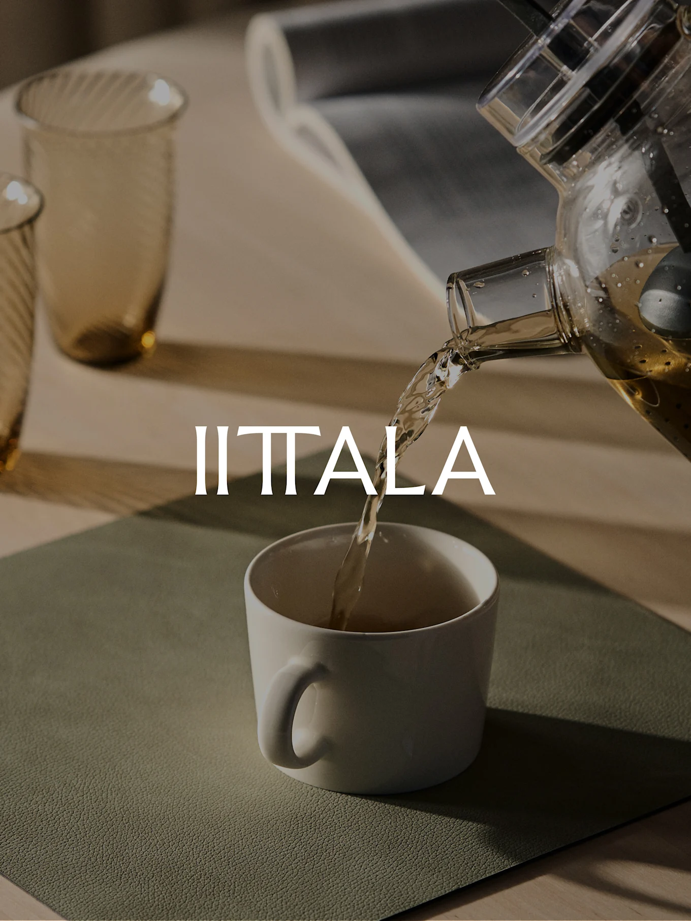 A clear teapot pours golden tea into a white mug, with two amber glasses and the Iittala logo.