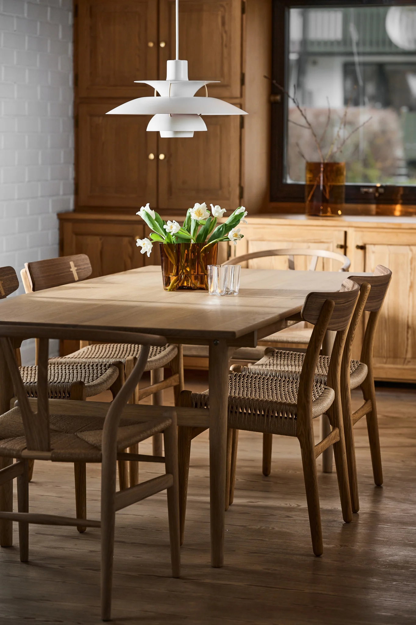 A modern dining room with a light wood table, woven-seat chairs, a white tiered lamp, and white flowers in a brown vase.