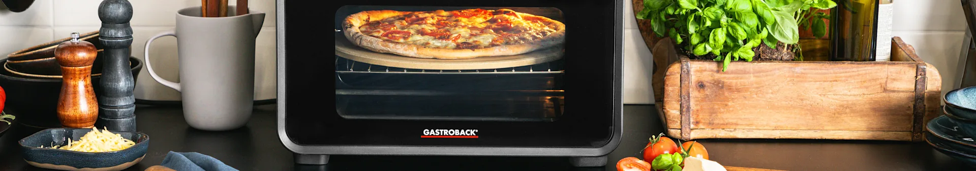 A GASTROBACK oven baking a cheesy pizza on a stone, with fresh basil, tomatoes, and grated cheese on the counter.