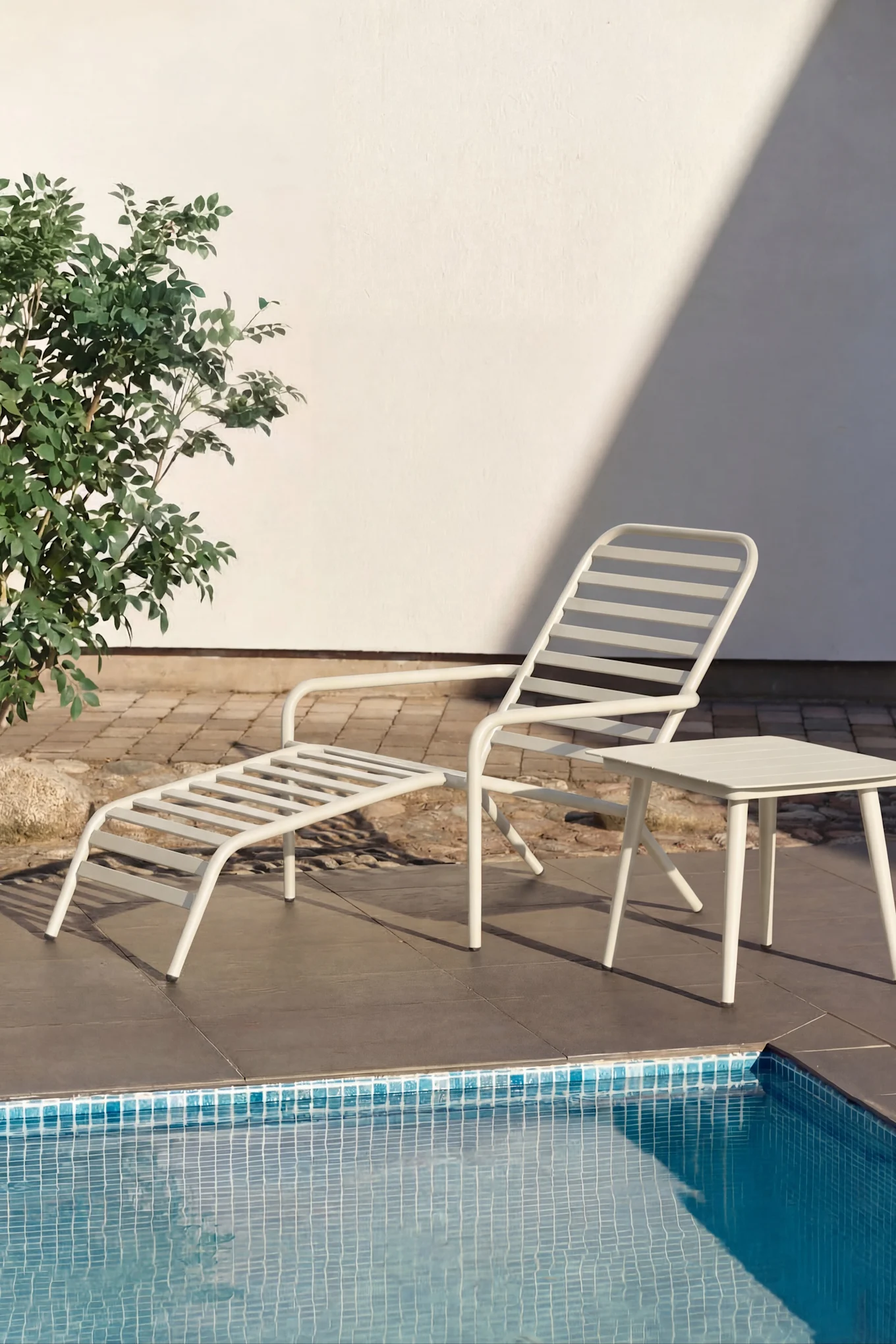 White lounge chair and small white side table by a pool with turquoise water. Green plants and a white wall in the background.