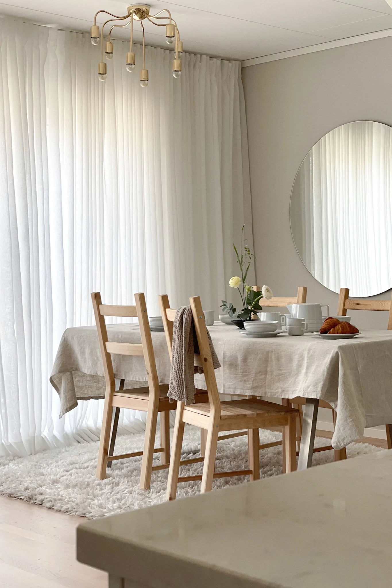 A bright dining room with a wooden table, white dishes, croissants, flowers, and wooden chairs.