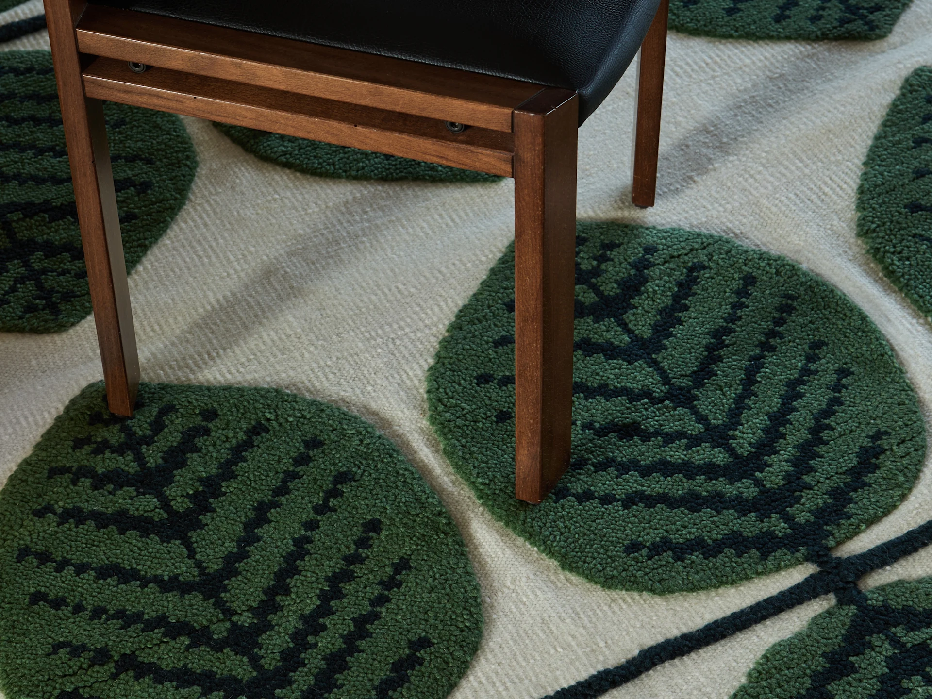 Dark brown wooden chair leg on a cream rug with a textured green leaf pattern.