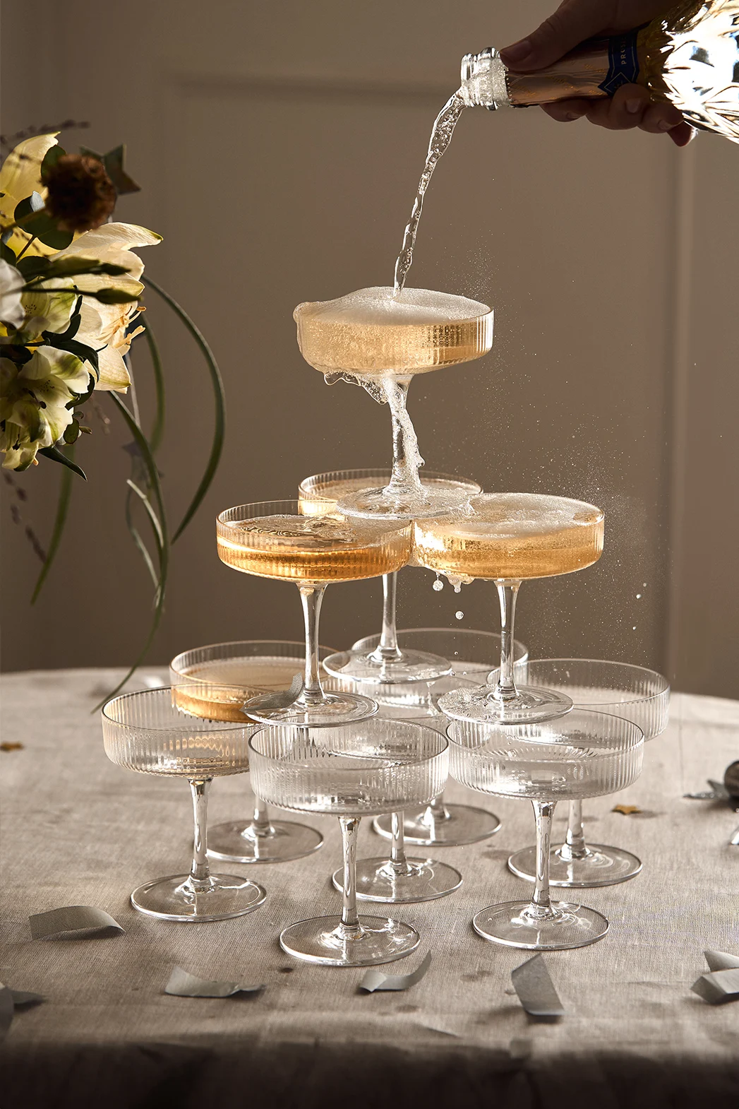 Champagne being poured from a bottle into a tower of ribbed coupe glasses, overflowing and cascading downwards.