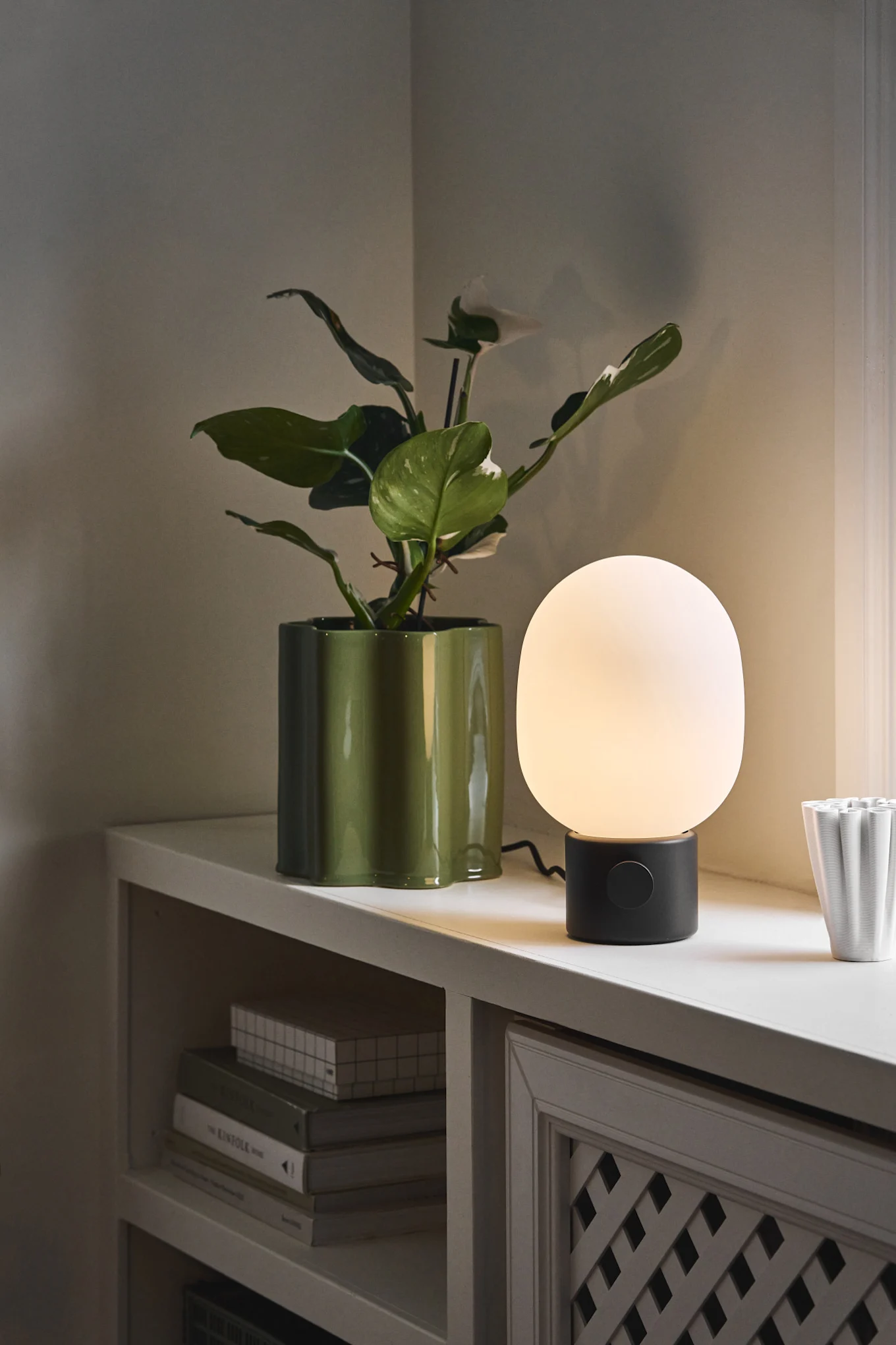 A glowing white table lamp with a black base next to a large plant in a glossy green wavy pot on a white shelf.