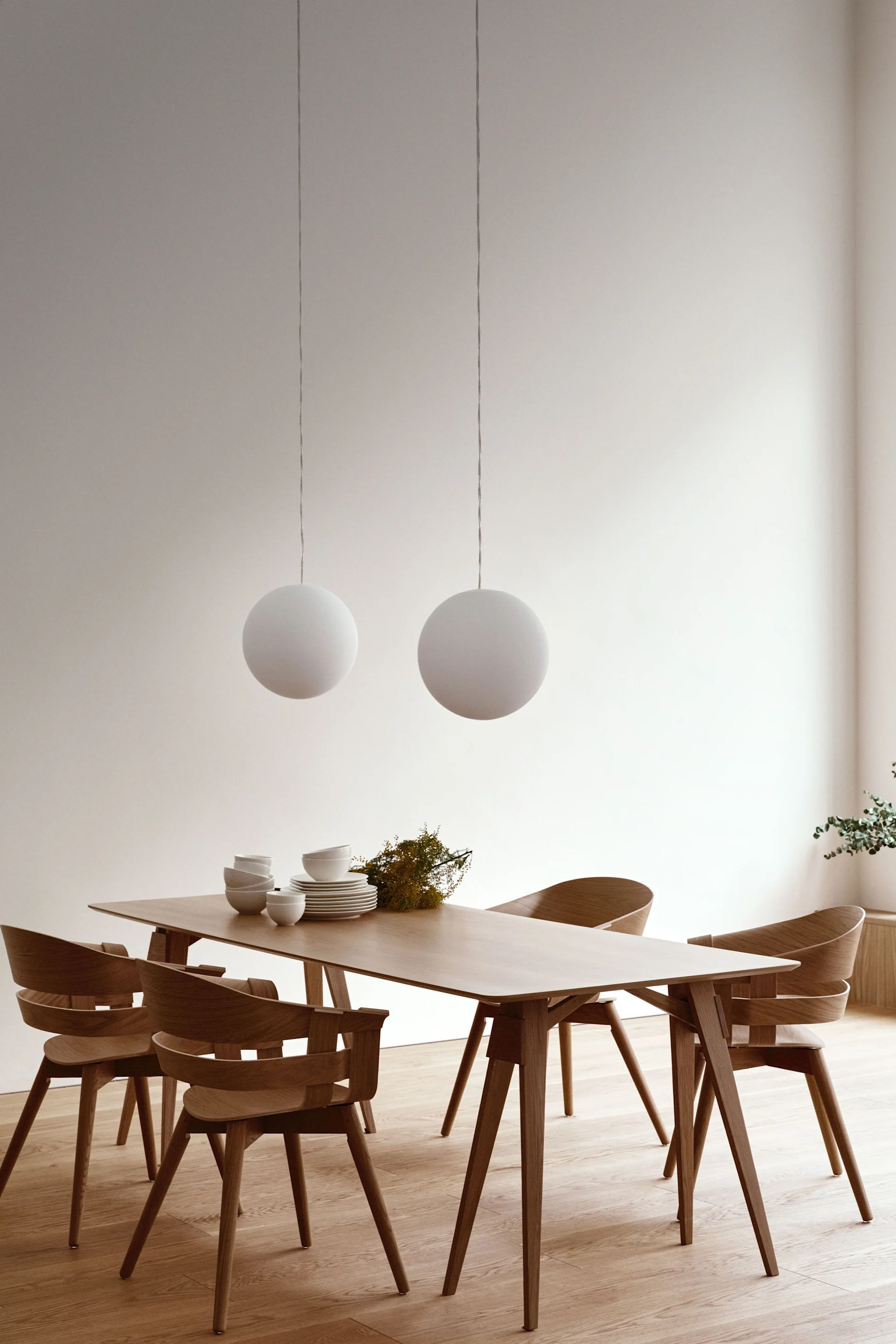 A modern dining room with a light wood rectangular table and four matching wood chairs. Two white spherical pendant lights hang above the table. White dinnerware and a small floral arrangement decorate the table. Light wood flooring and white walls complete the minimalist aesthetic.