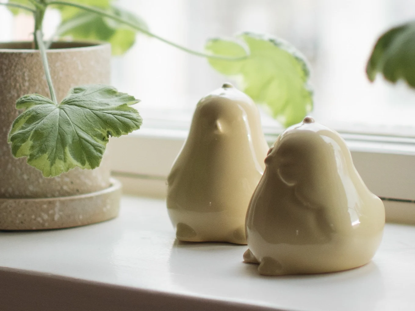 Two glossy, light yellow ceramic bird figurines sit on a white windowsill next to a potted plant with variegated leaves.