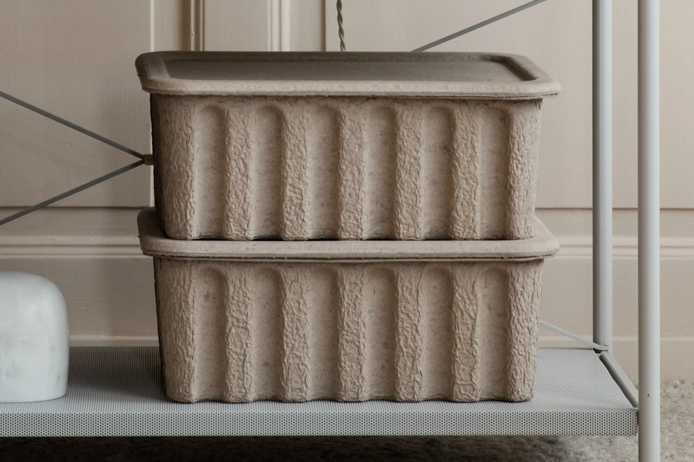 Two stacked light brown rectangular storage boxes with vertical ridges, on a light grey perforated metal shelf.