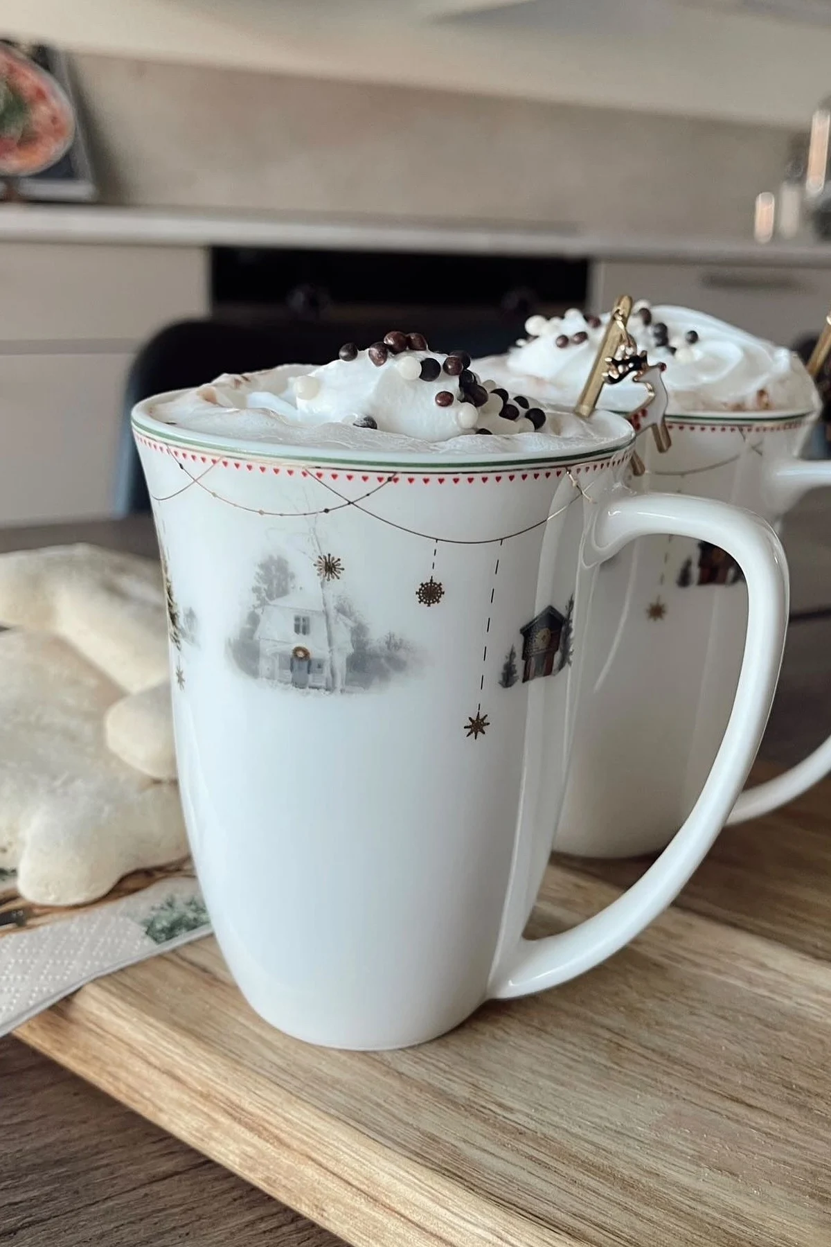 Two white mugs with festive winter scenes, topped with whipped cream, chocolate sprinkles, and a reindeer spoon, next to star cookies.