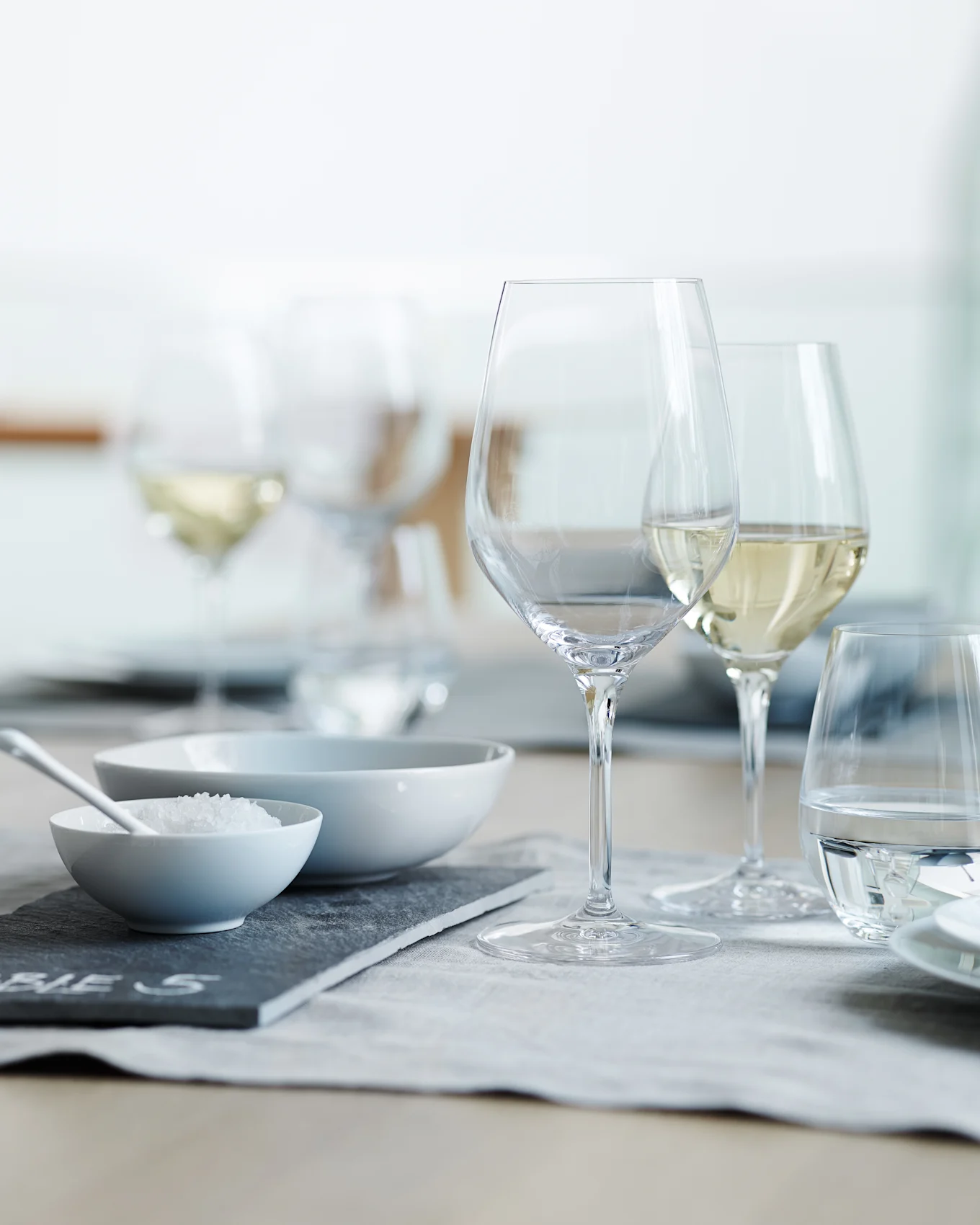 A dining table set with clear wine and water glasses, a bowl of coarse salt, and an empty bowl on a grey placemat.