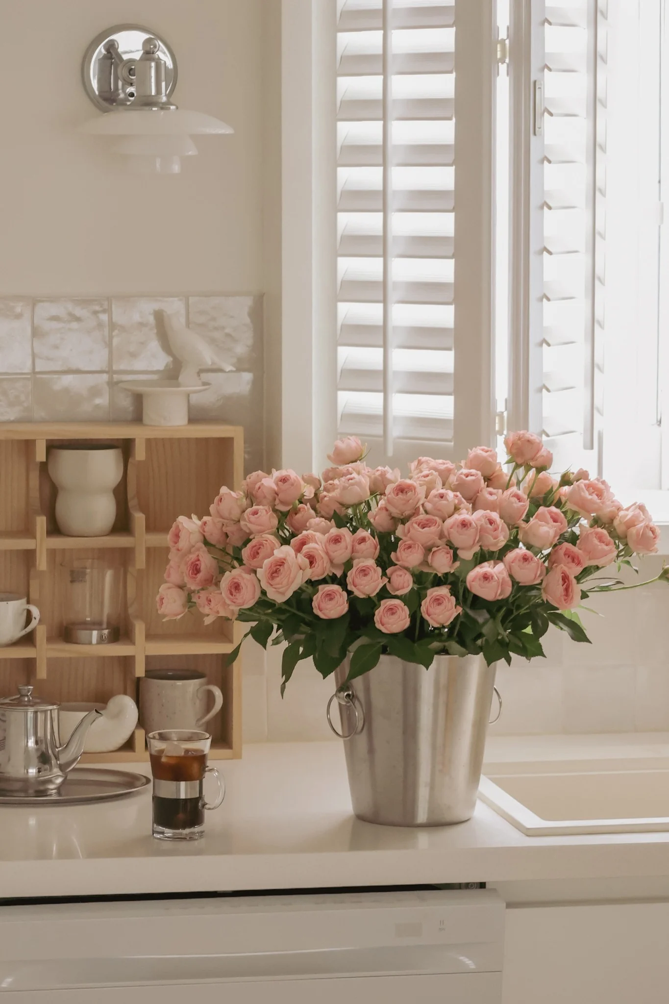 A large bouquet of pink roses in a silver bucket sits on a white kitchen counter next to a window with shutters.