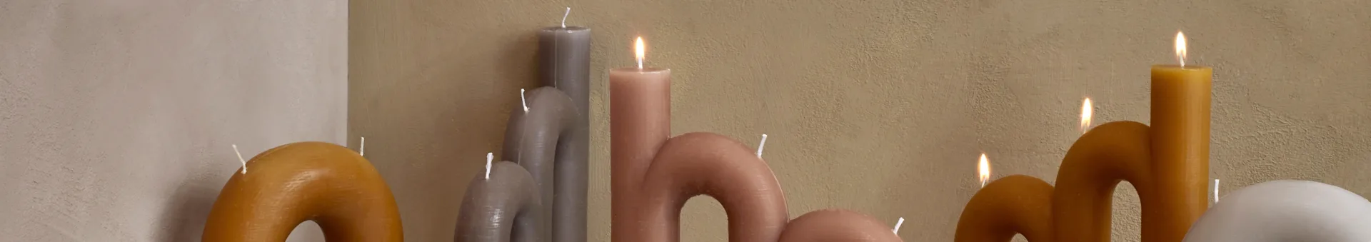 A collection of wavy, curved candles in shades of yellow, grey, and pink, with some lit, against a textured wall.