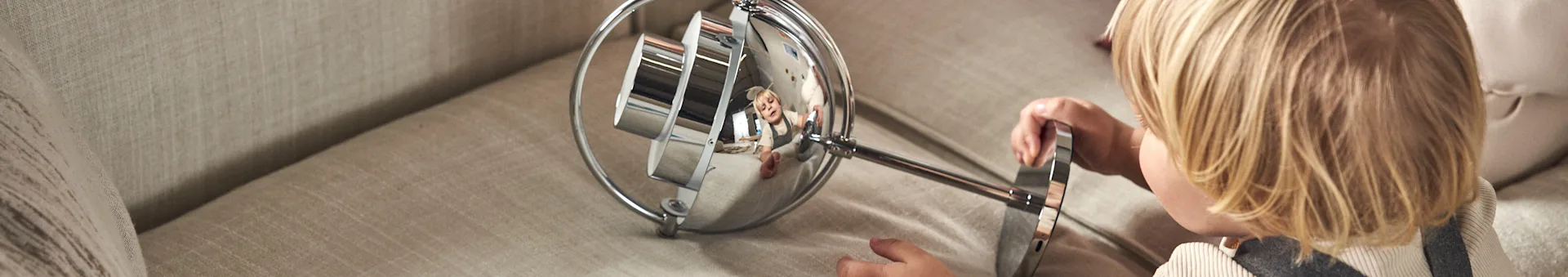 Child with blonde hair looking into a chrome mirror that reflects a smiling adult.