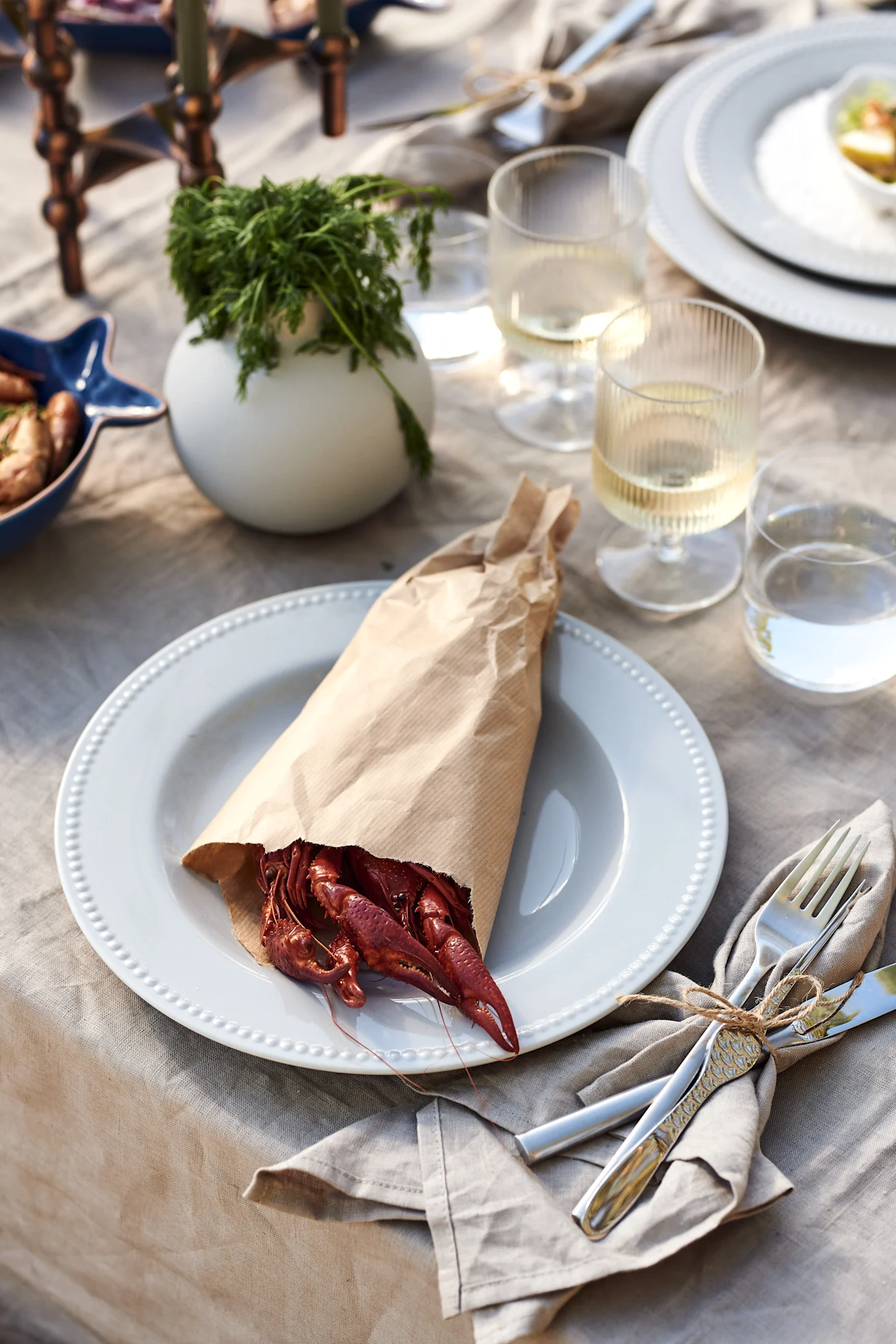 Cooked red crayfish spilling from a paper bag onto a white plate on an outdoor table set with cutlery and drinks.