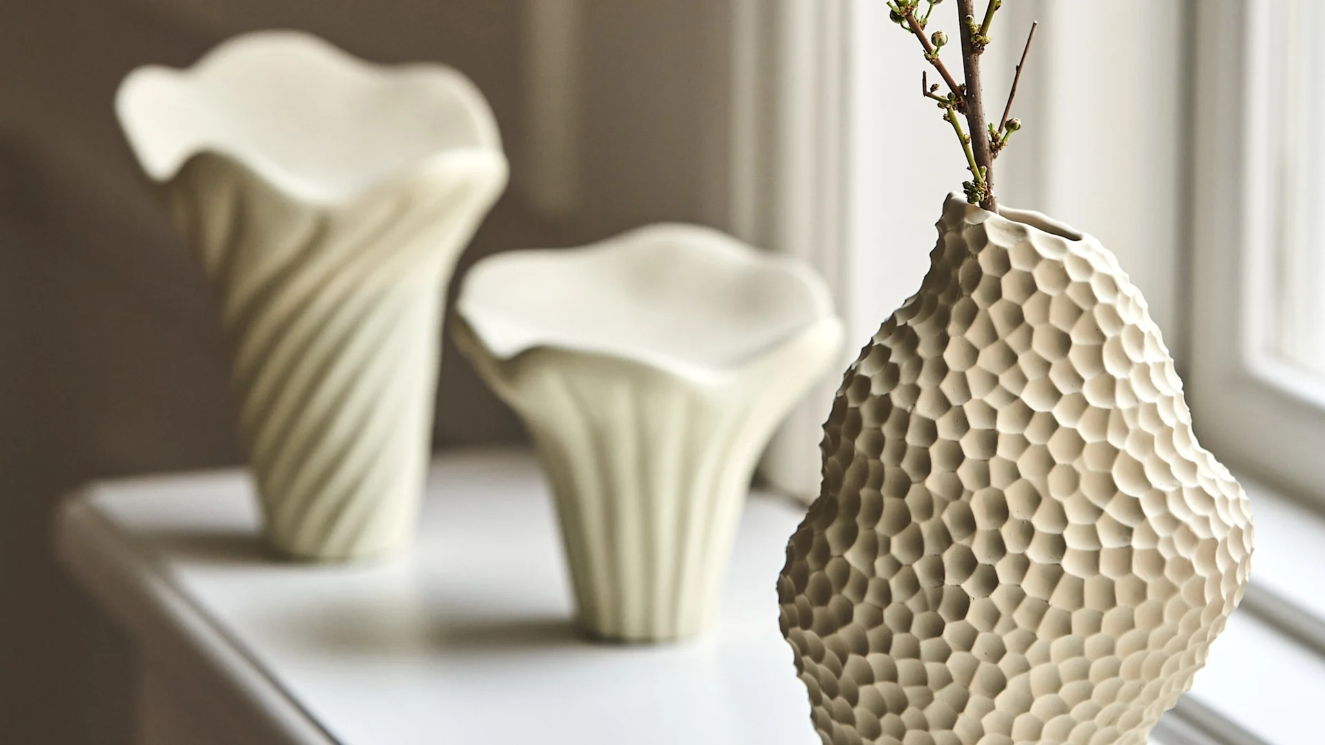 White hexagonal textured vase with a budding branch, next to two organic-shaped vases on a white shelf.