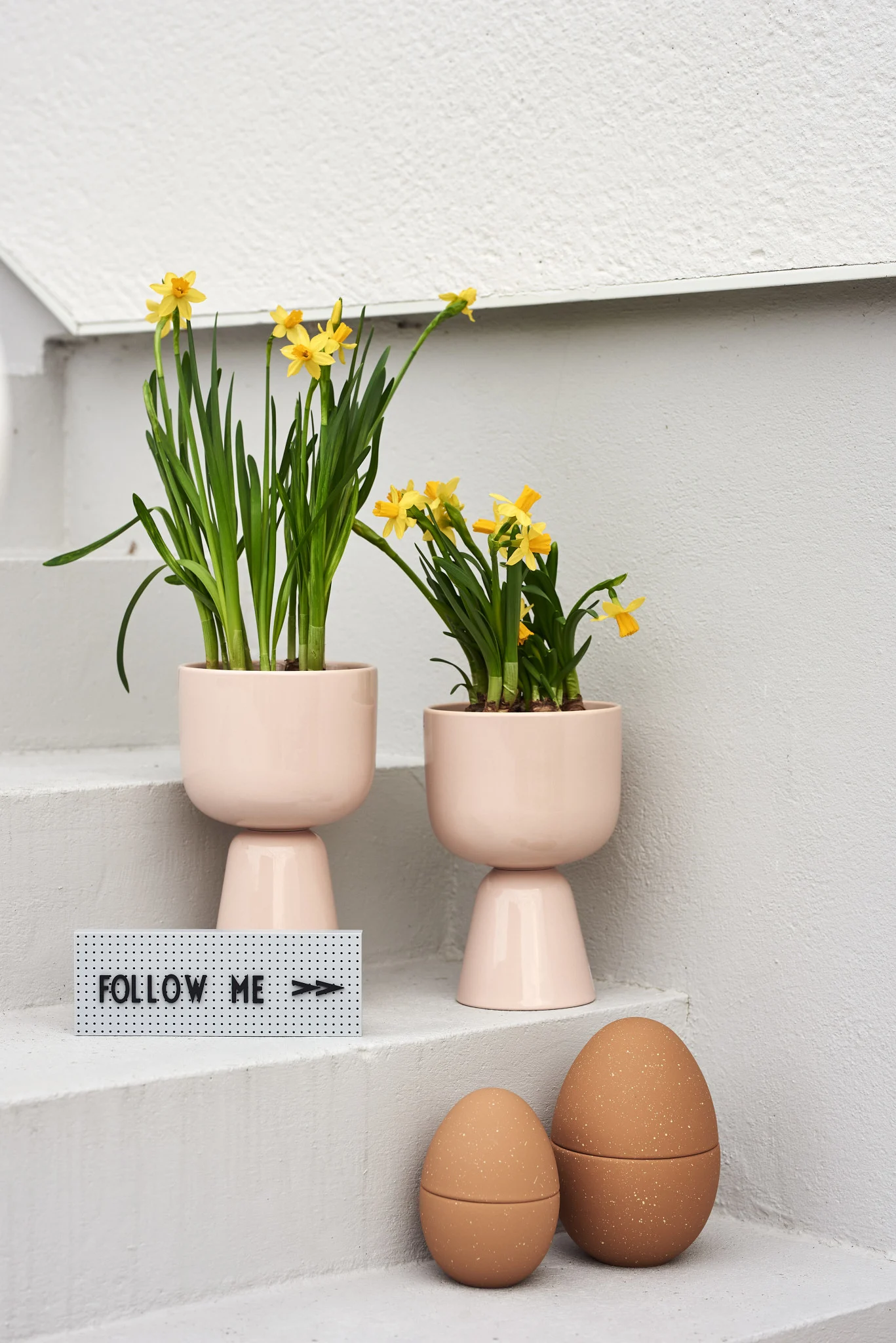 Two pink planters with yellow daffodils on white steps, a 'FOLLOW ME' sign, and two speckled brown decorative eggs.