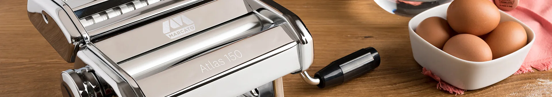 Chrome pasta maker, bowl of eggs, and flour on a wooden table.