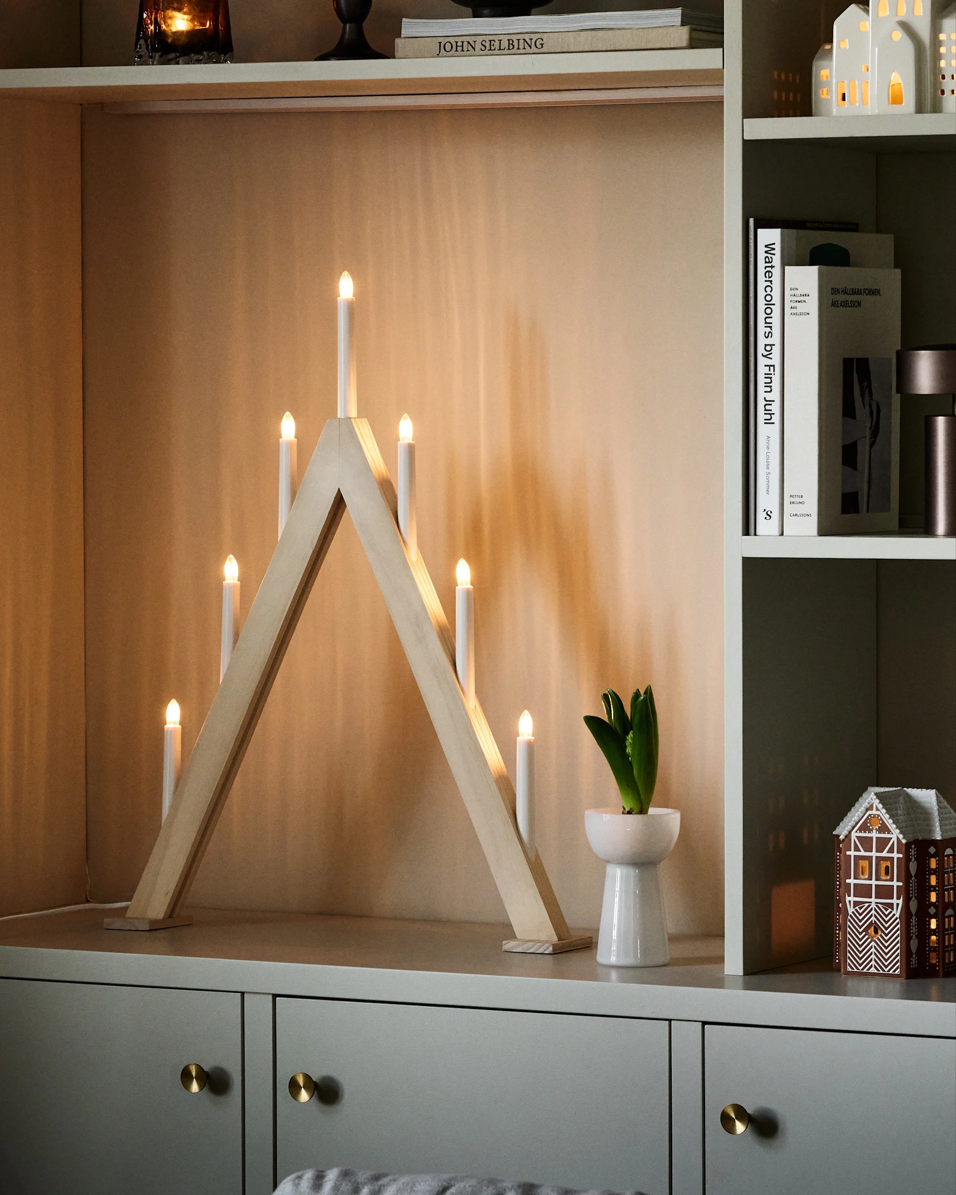 A wooden candle arch with seven electric candles and a potted hyacinth on a cabinet, with decorative houses on shelves.