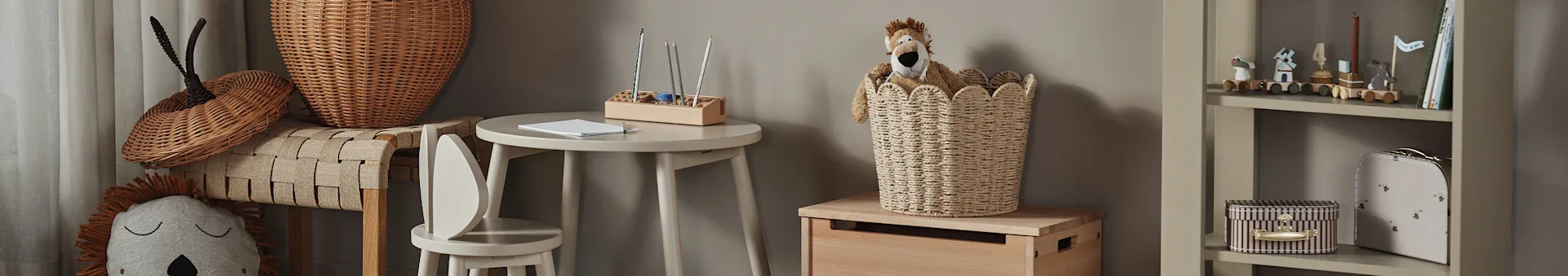 A child's room with a lion plush toy in a woven basket, a small wooden table with pencils, and a bookshelf with toys and books.