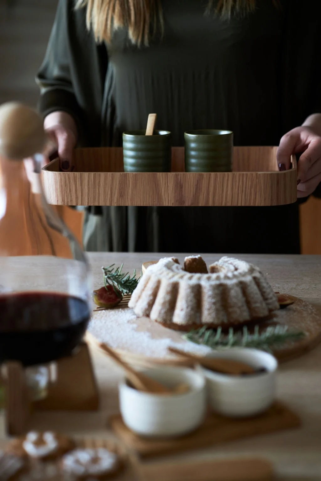 Person in green holds wooden tray with two dark green ribbed cups. Bundt cake, rosemary, fig, and red beverage are on the table.