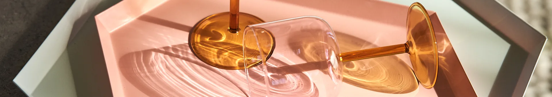 Two wine glasses with amber stems on a pink rectangular tray inside a white hexagonal tray, with sunlight creating patterns.