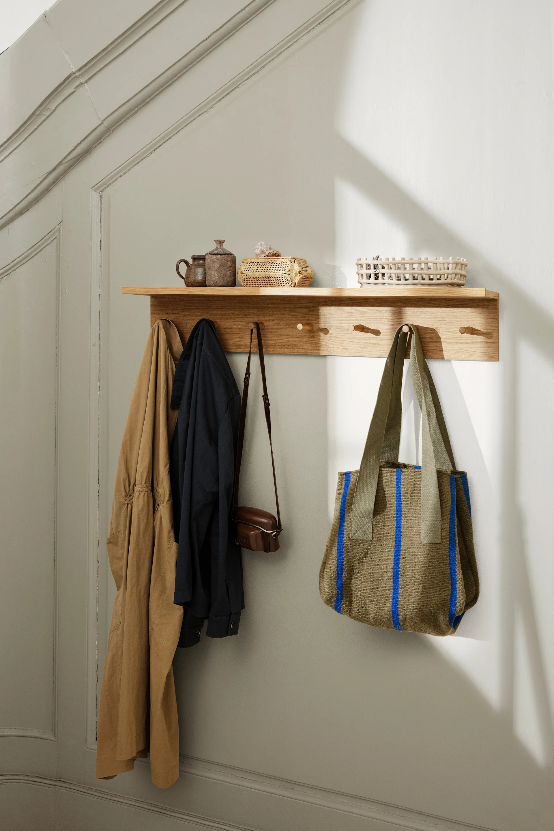 A wooden wall shelf with pegs holds two jackets, a small leather bag, and a large olive green tote with blue stripes, above which sit decorative pots and baskets.