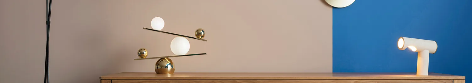 A modern gold balance lamp with white globes and gold spheres on thin rods, next to a white cylindrical lamp with an illuminated opening, on a wooden surface against a two-tone beige and blue wall.