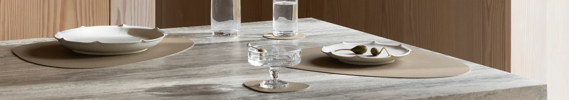 A marble table set with two scalloped bowls, three glasses, and ribbed beige placemats against a wooden background.