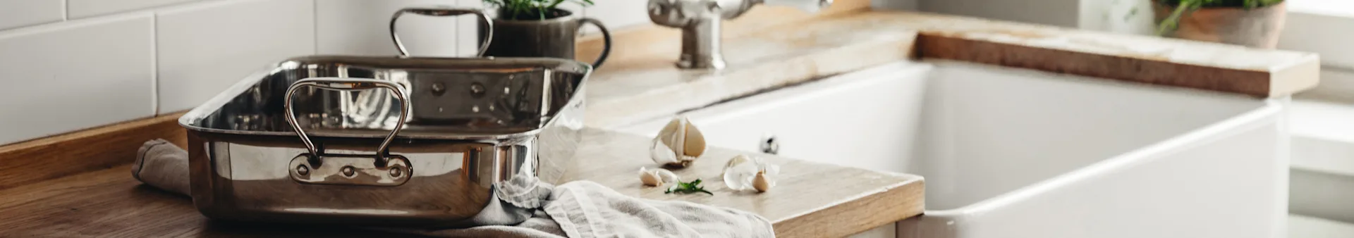 A stainless steel roasting pan, garlic, and herbs on a wooden kitchen counter next to a white farmhouse sink.