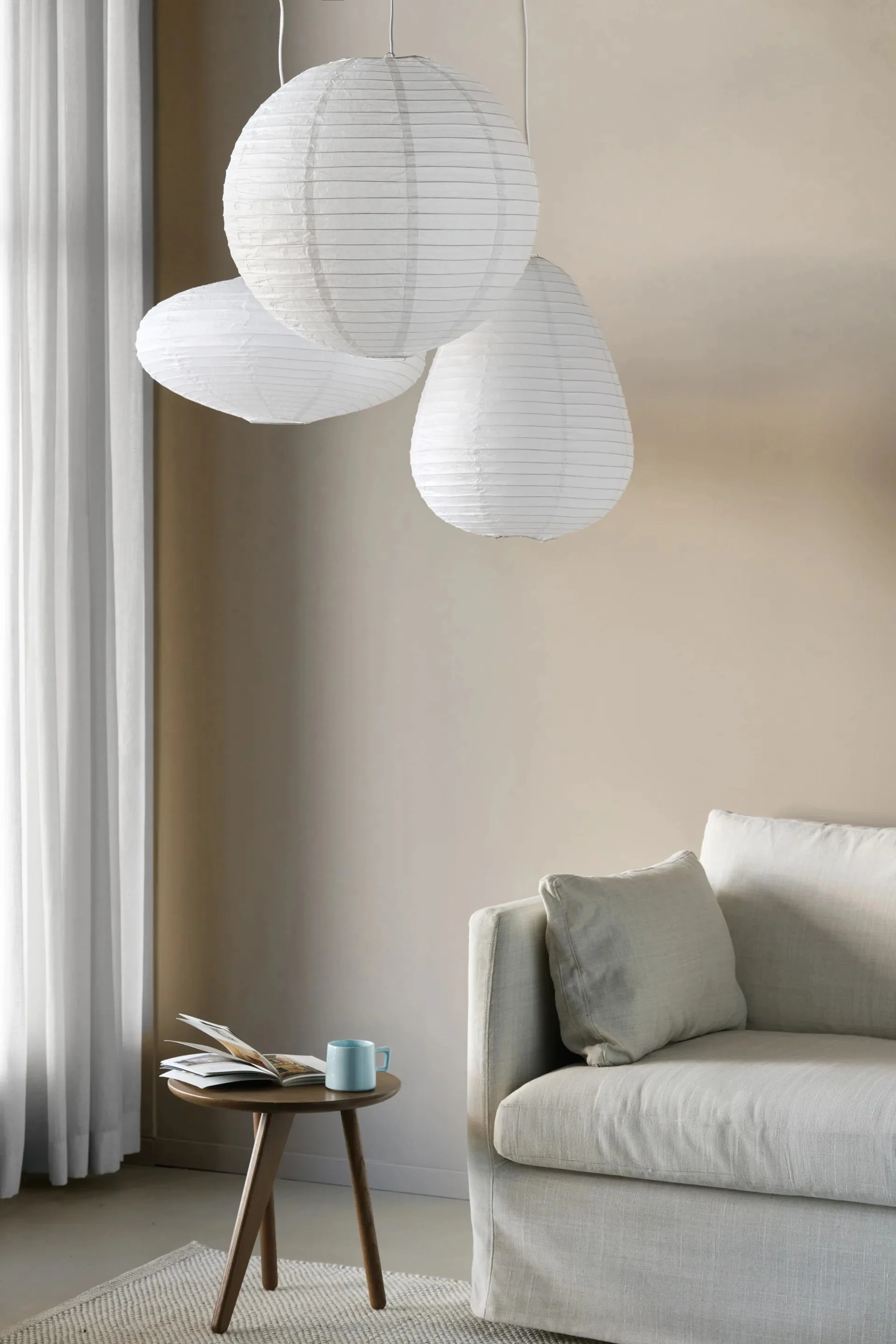 Three white paper lanterns hanging above a neutral-colored sofa, a wooden side table with a book and a blue mug, and white curtains.