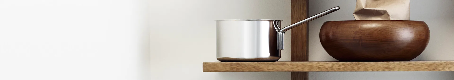 A shiny silver saucepan and a dark wooden bowl with a paper bag on a light wooden shelf.