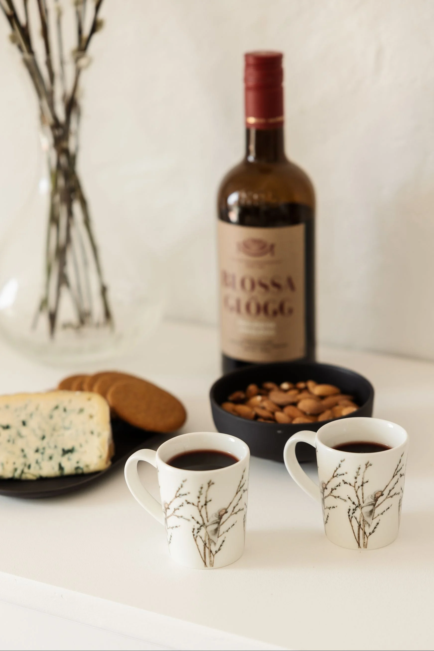 Two white mugs with branch design, filled with mulled wine, a bottle of Blossa Glögg, blue cheese, cookies, and almonds.