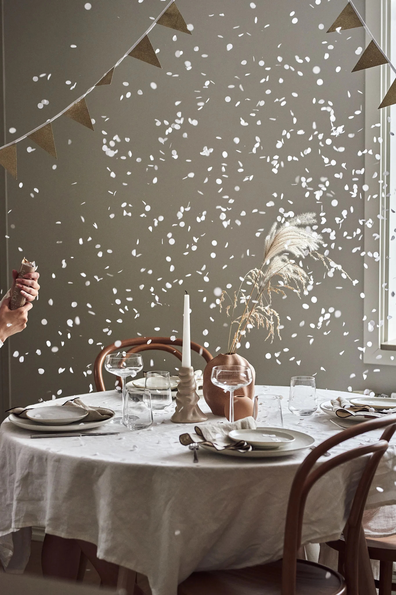 Hands pop confetti over a table set for a celebration, with plates, glasses, a vase with pampas grass, and a candle.