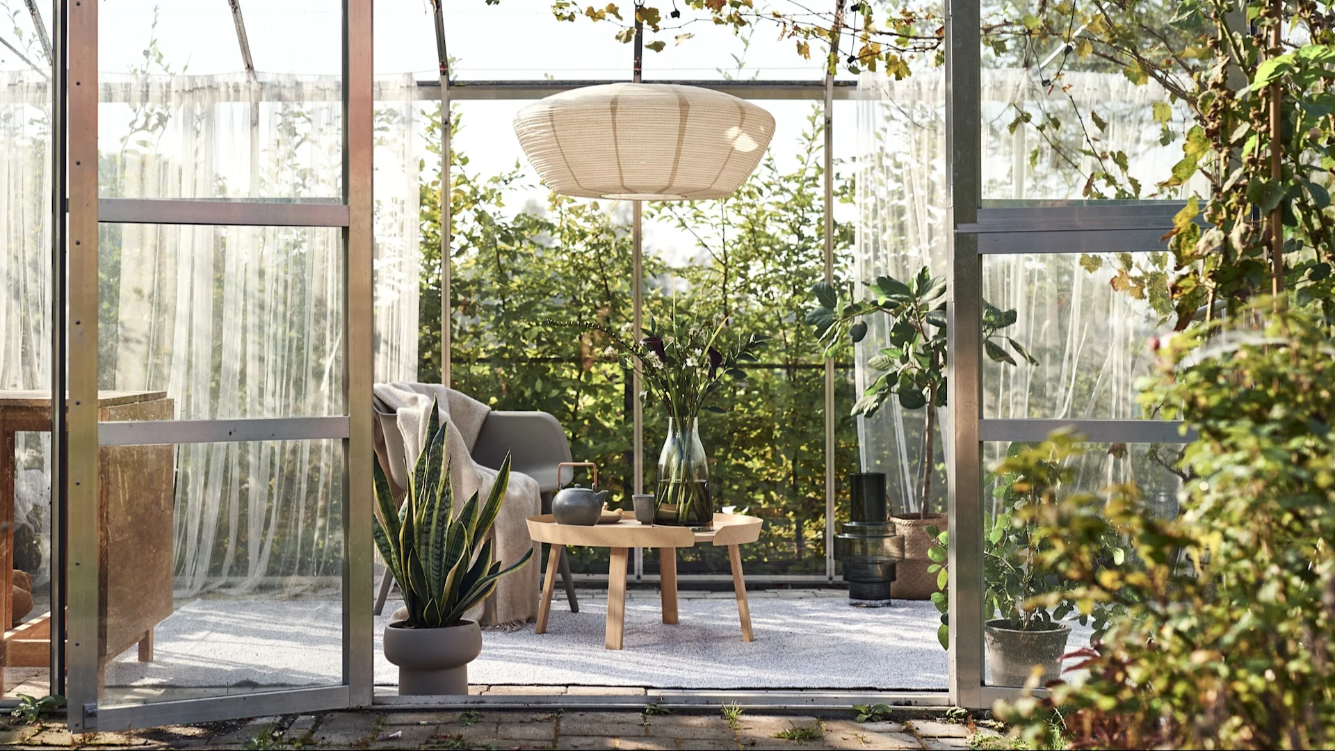 Cozy greenhouse interior with pendant lamp, wood table, armchair, snake plant, and vase with flowers.