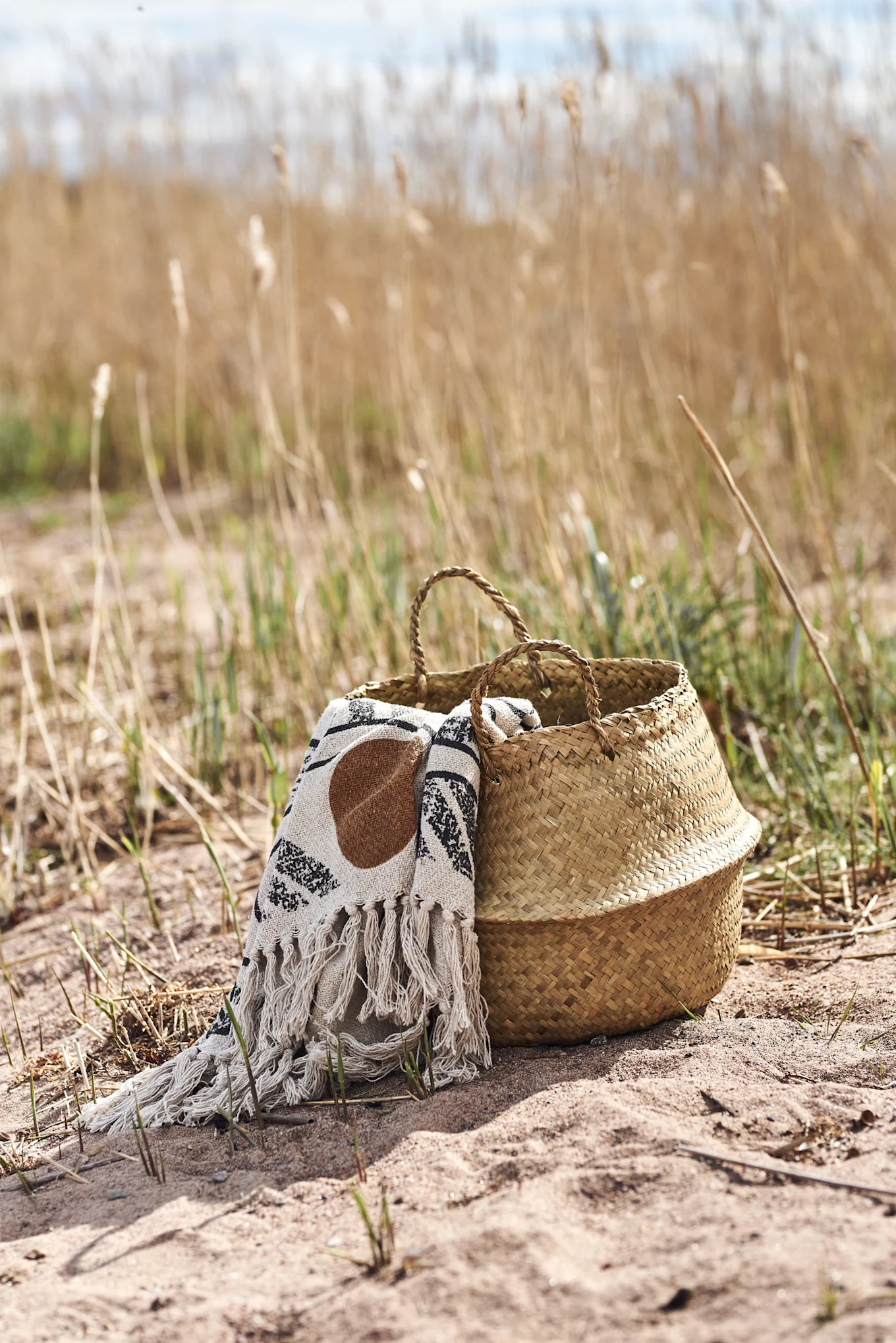 A woven straw basket with a white patterned blanket on sand, surrounded by dry reeds.