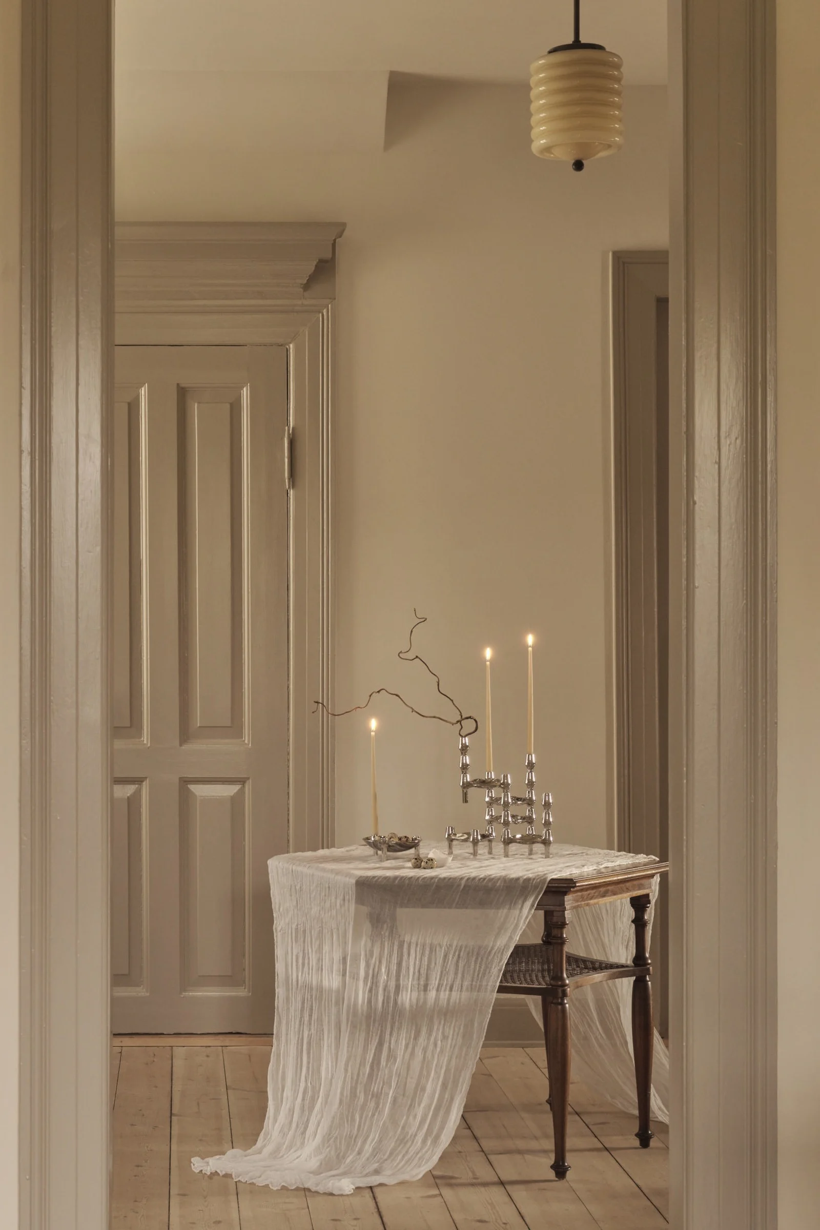 A wooden table with a white sheer cloth holds three lit candles in silver candelabras, a bowl of speckled eggs, and a branch in a muted room.